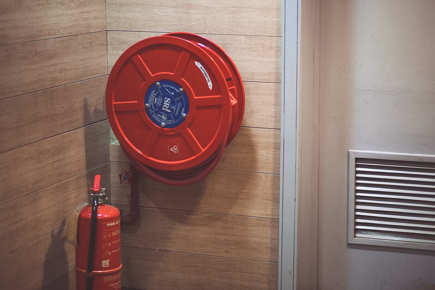 Red fire hose reel mounted on a wood-paneled wall beside a doorway, with a red fire extinguisher standing in the corner below