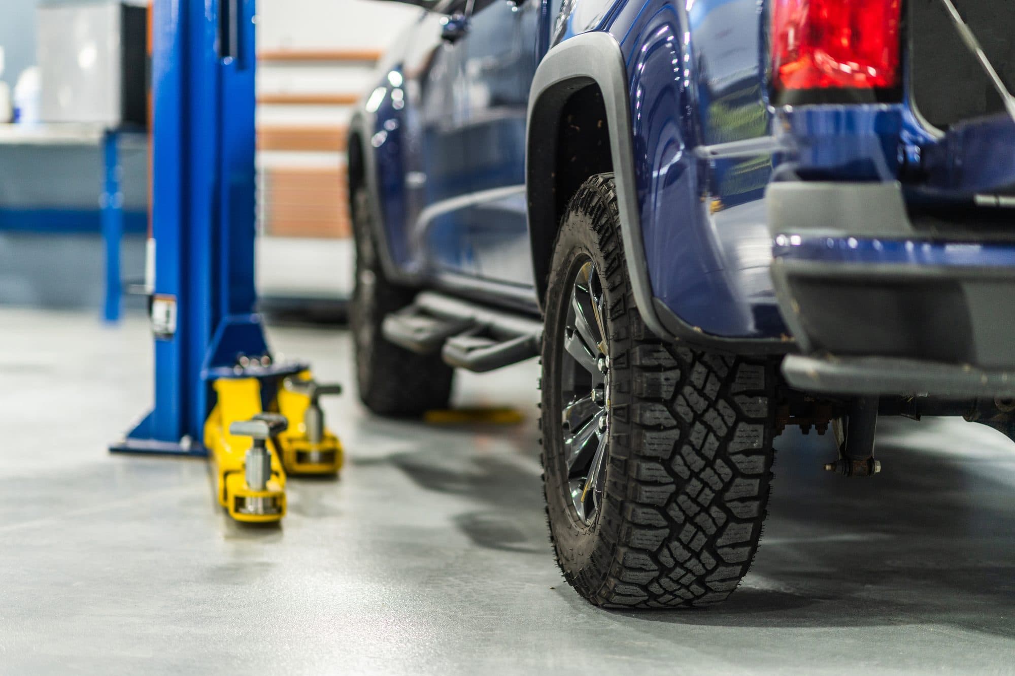 Blue pickup truck parked in an automotive service bay, photographed from floor level, with a two-post lift column and a yellow hydraulic floor jack behind it on a grey concrete shop floor