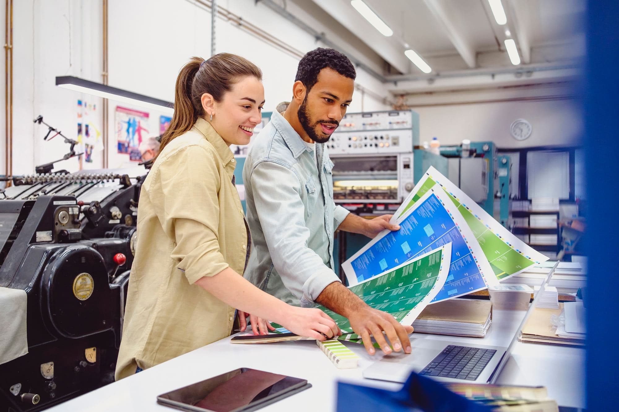 Two coworkers reviewing printed press sheets together beside a press in a commercial printing facility