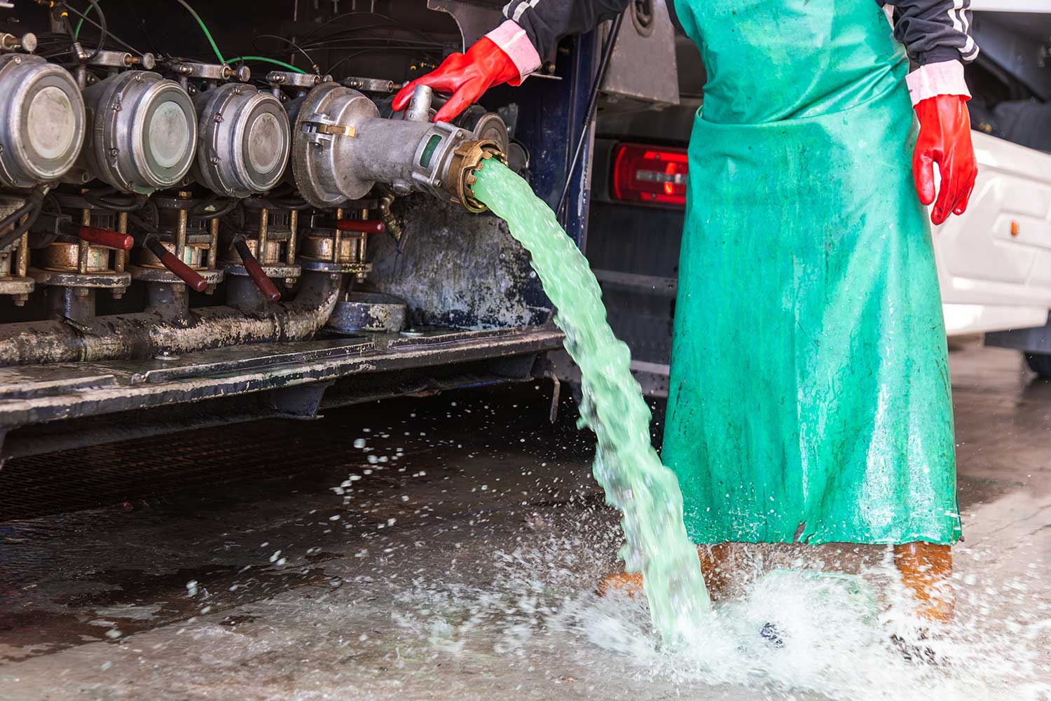 Worker in a green apron and red rubber gloves draining green liquid from a tanker truck outlet, the fluid splashing onto the pavement