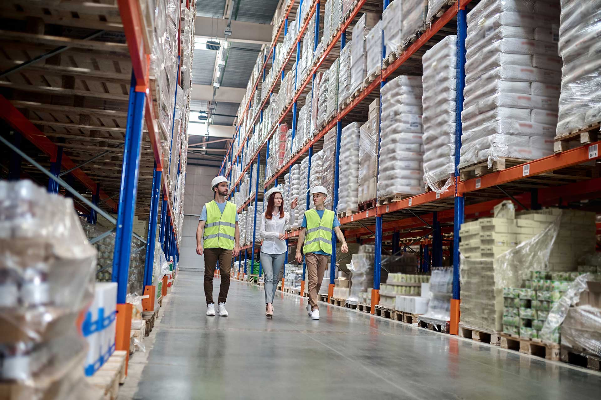 Three people in white hard hats and hi-vis vests walking down a tall warehouse aisle between racks of shrink-wrapped pallets