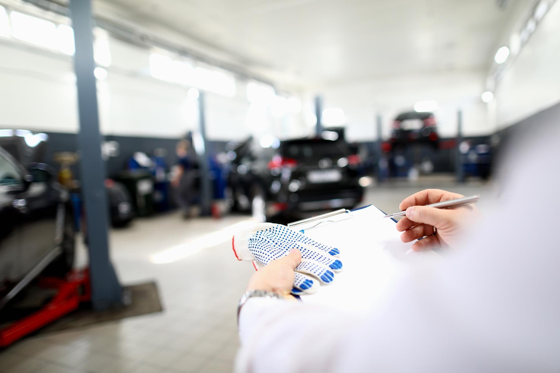 Inspector holding a clipboard, pen, and knitted work gloves in a service garage with cars raised on lifts in the background