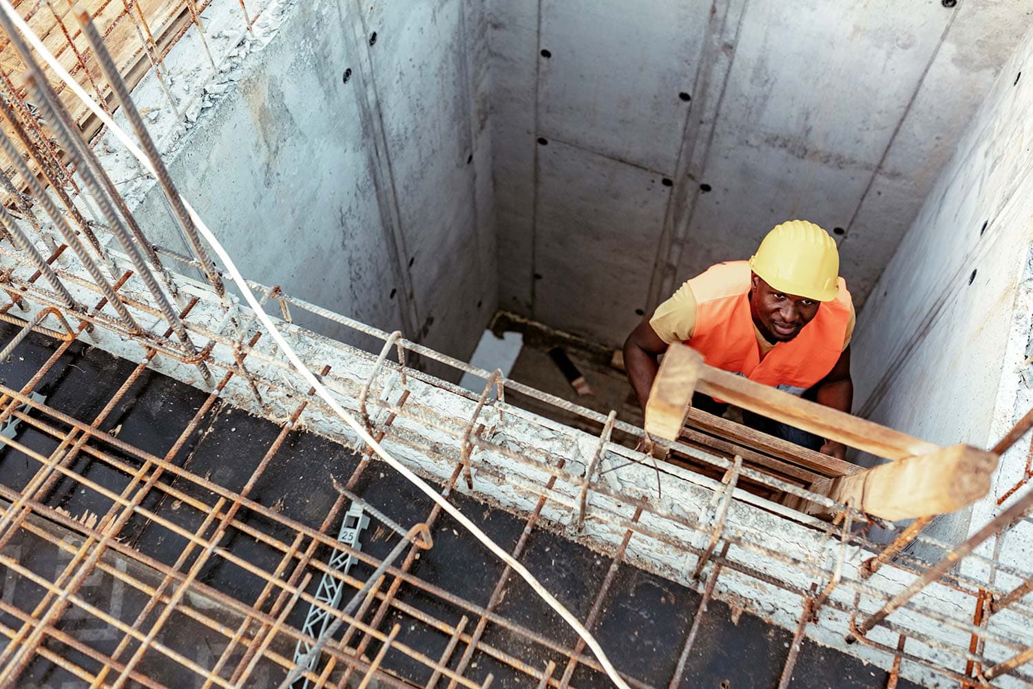 Worker in a yellow hard hat and orange vest climbing a wooden ladder out of a deep concrete shaft edged with rebar, seen from above