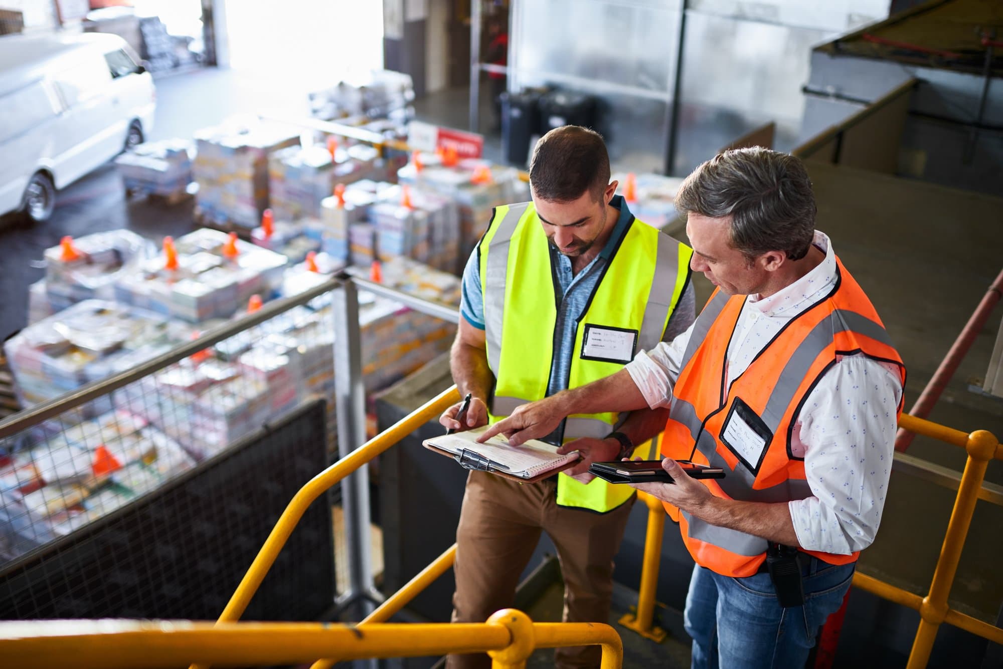 Two workers in high-visibility safety vests reviewing a clipboard on an elevated industrial walkway.