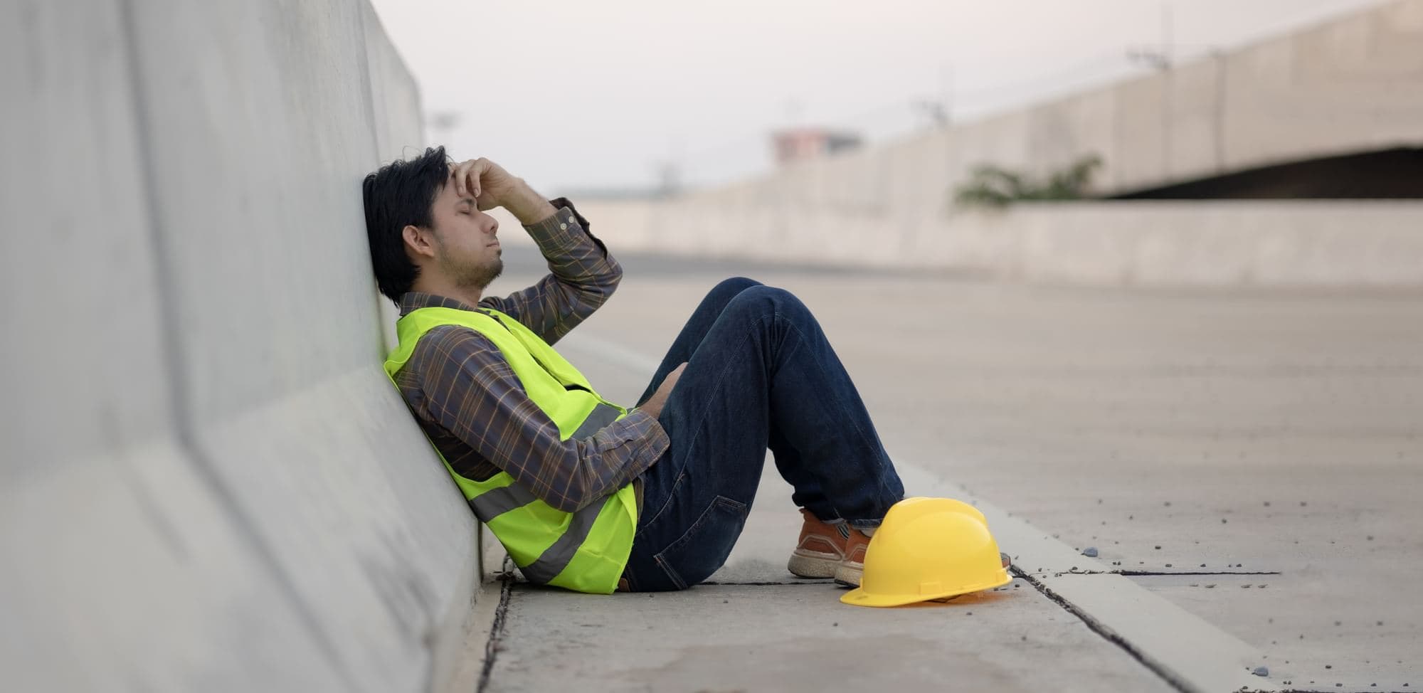 Construction worker in a high-visibility vest sitting beside a concrete barrier with a hard hat on the ground.