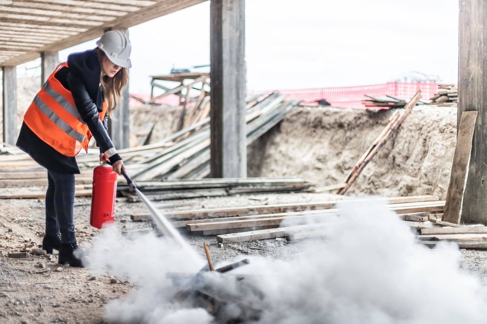 Worker wearing a hard hat and high-visibility vest using a fire extinguisher at a construction site.