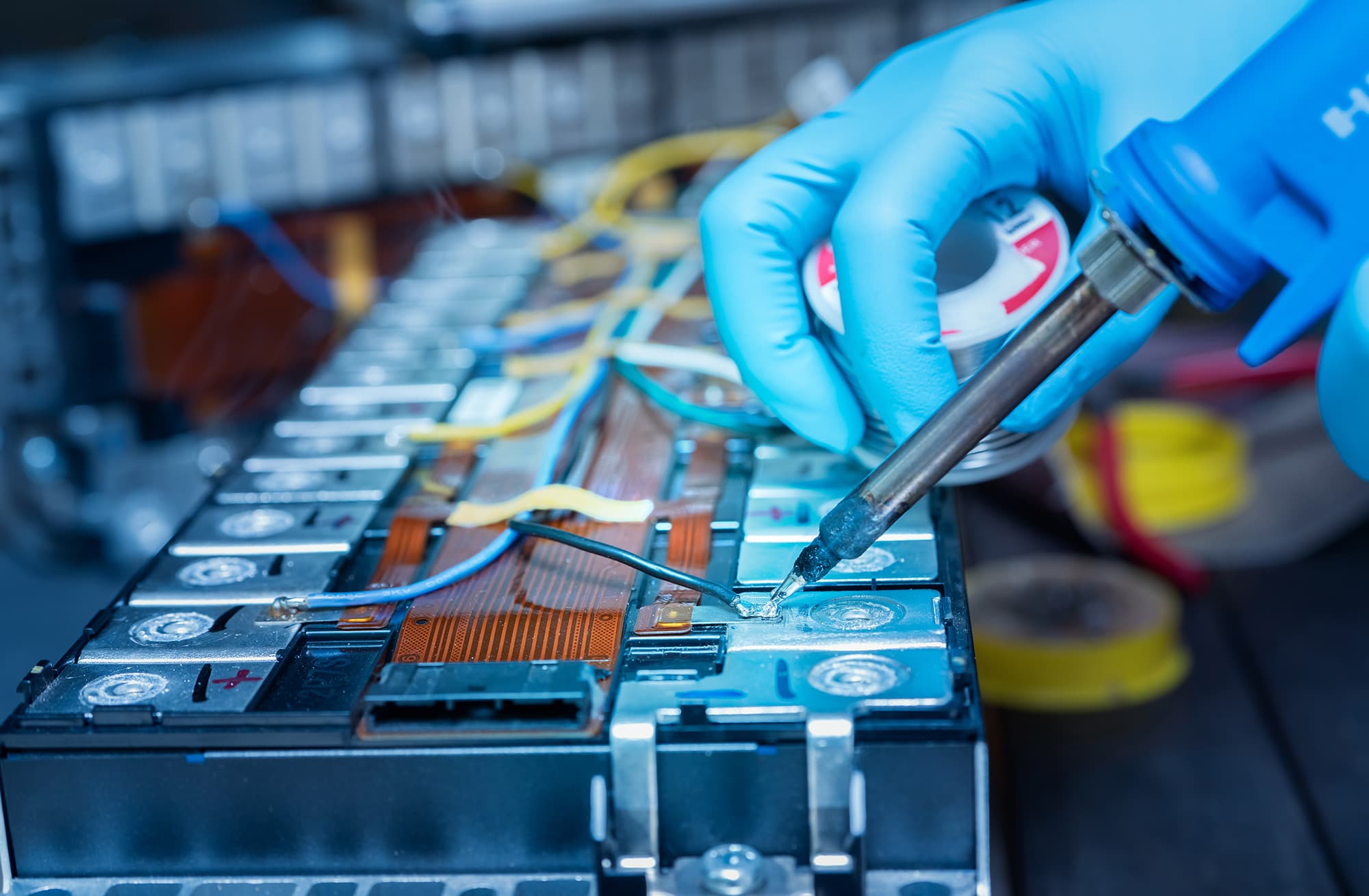 Technician in blue gloves soldering a wire on an electric vehicle battery module while holding a spool of solder