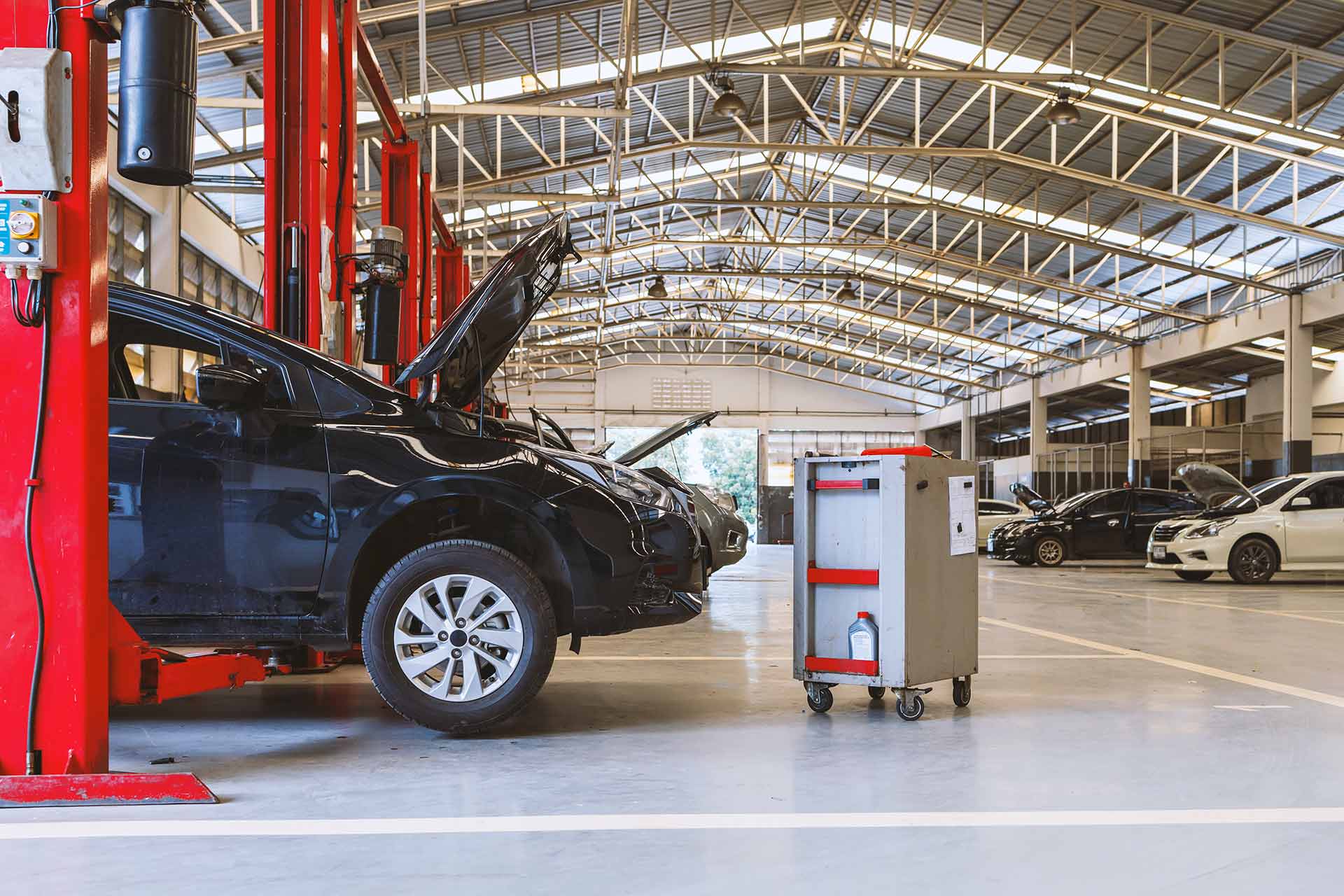 Black car with its hood open on a red two-post lift in a spacious service garage, a wheeled equipment cart parked beside it