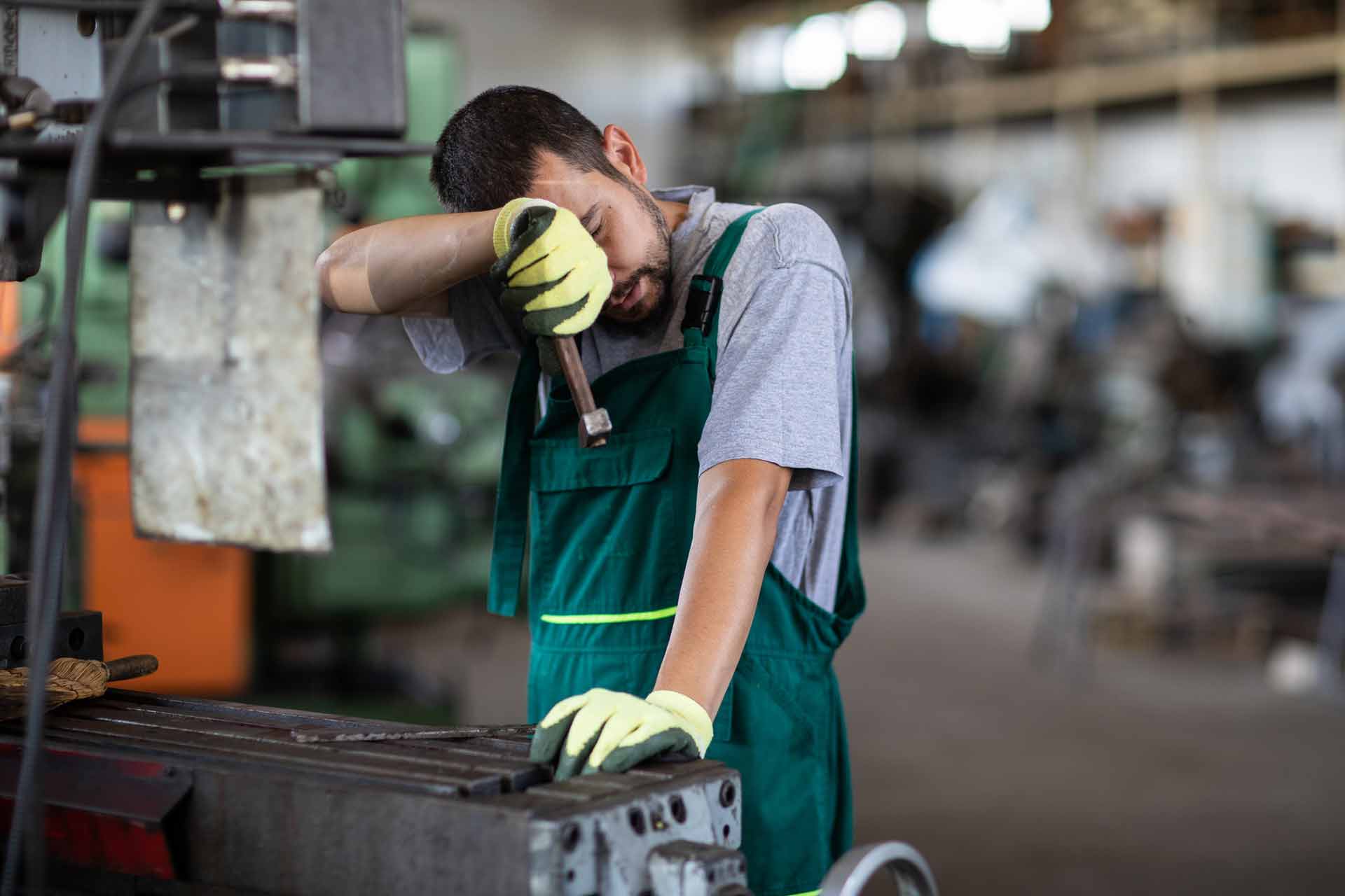 Weary machine operator in green overalls and work gloves wiping his brow with his forearm while leaning on shop equipment
