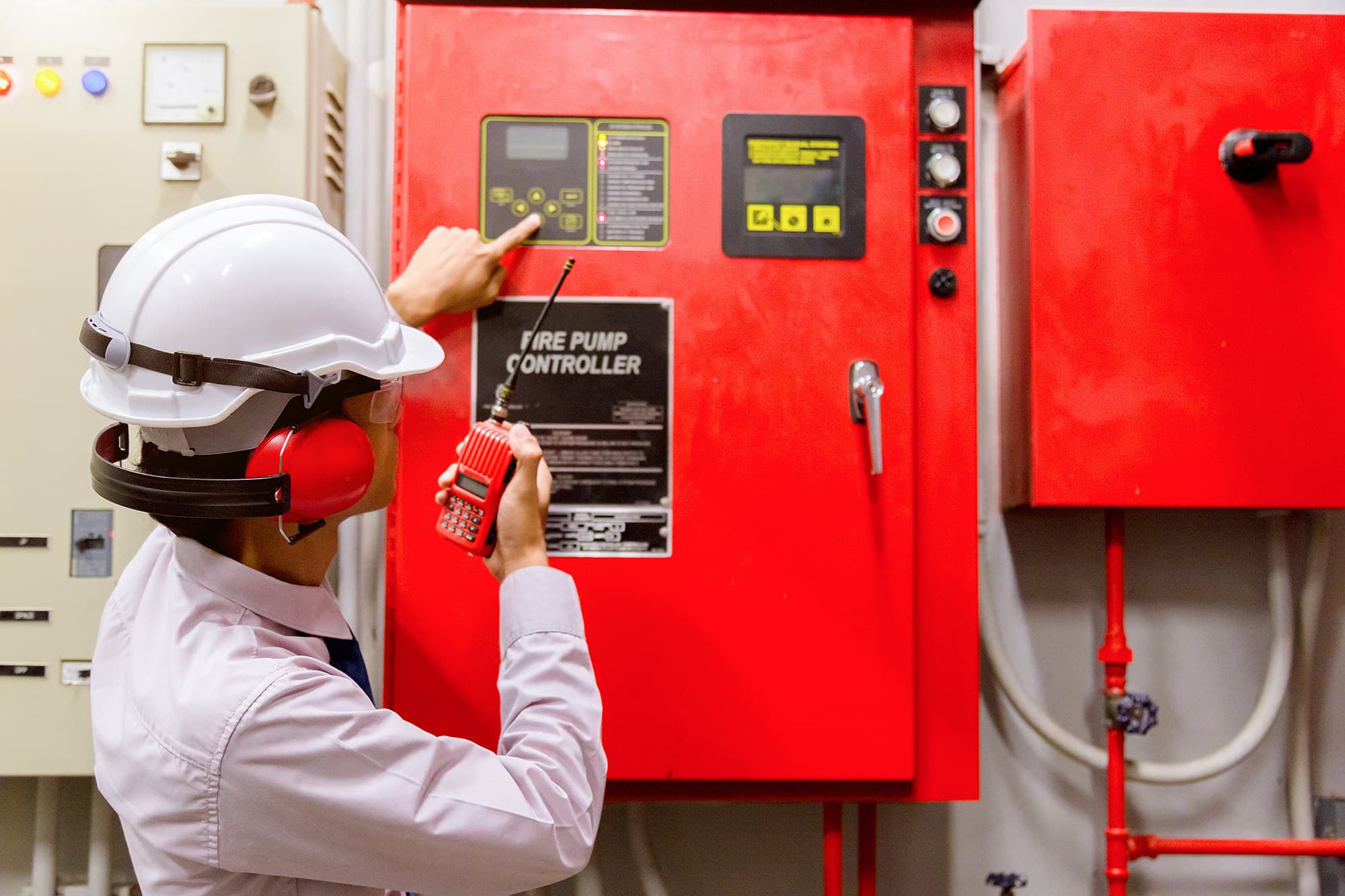 Worker in a white hard hat and red ear muffs holding a red two-way radio and pointing at a wall-mounted red fire pump controller panel