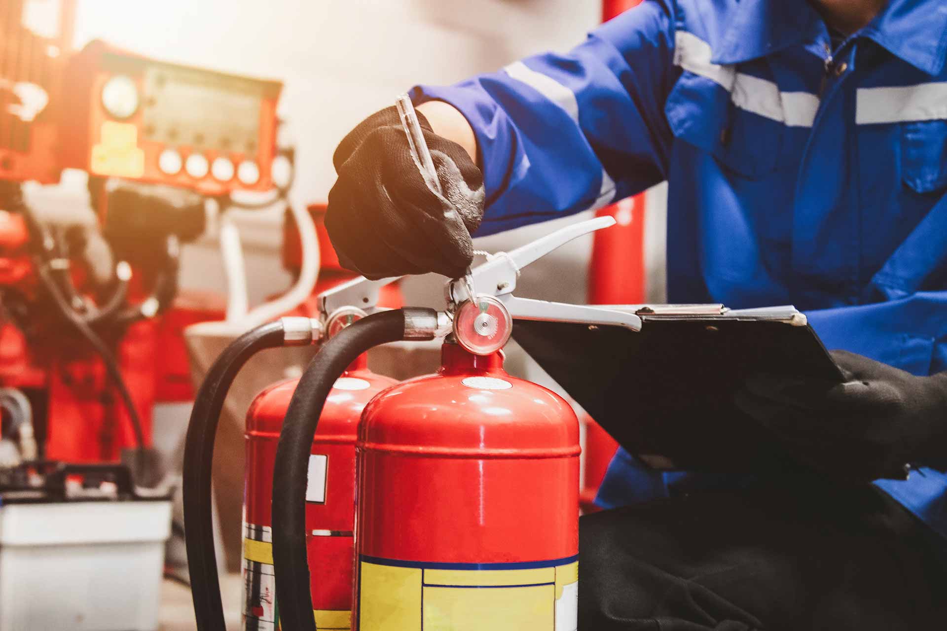 Worker in blue coveralls with a clipboard checking the pressure gauge on top of two red fire extinguishers