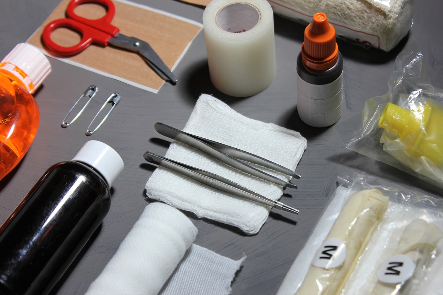 Flat lay of first aid supplies on a gray table, including bandage scissors, tweezers, gauze pads, tape, safety pins and antiseptic bottles