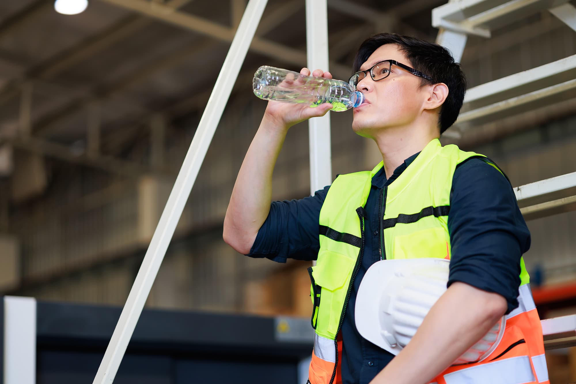 Warehouse worker in a yellow high-visibility vest and glasses drinking from a water bottle while holding a white hard hat