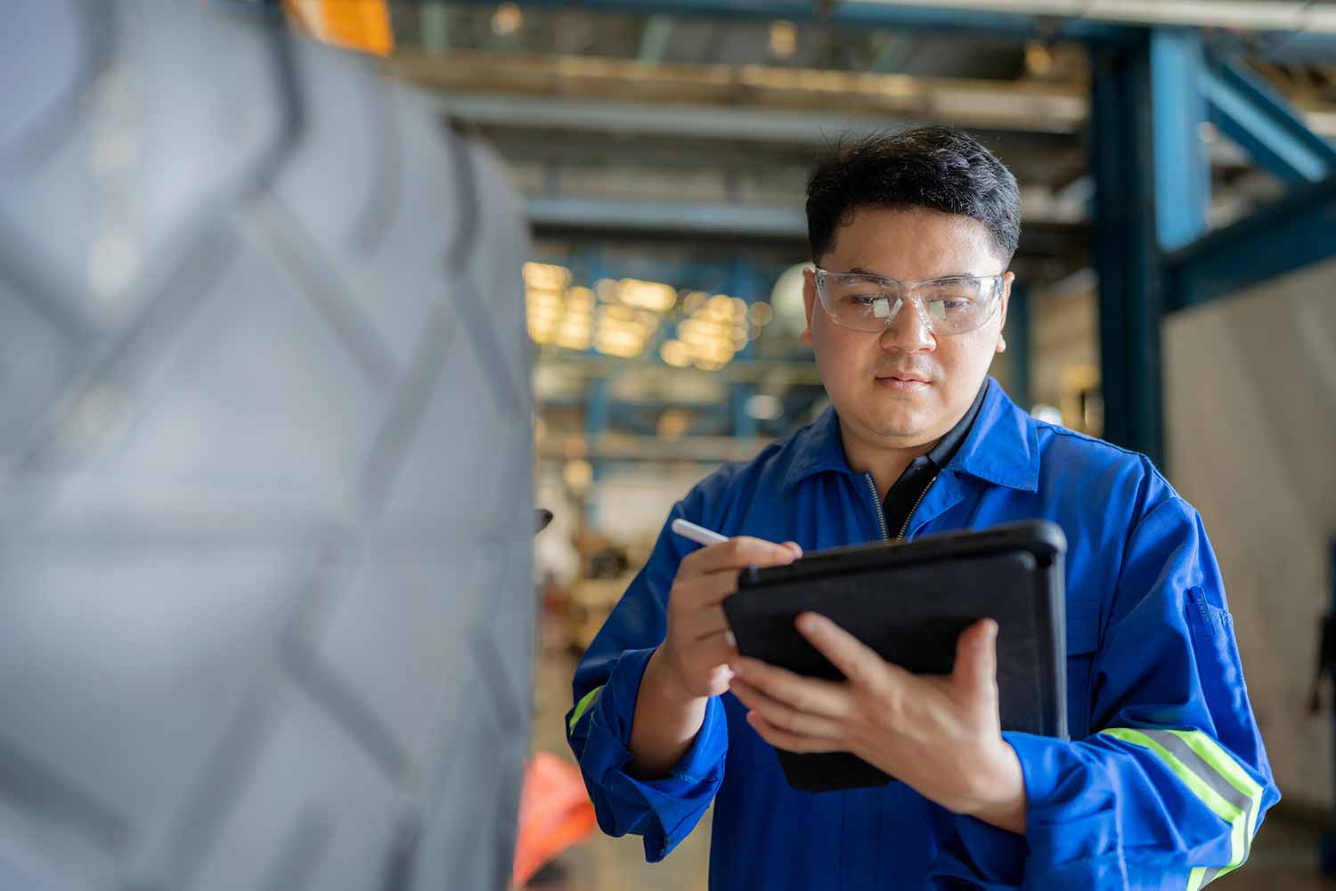 Technician in blue coveralls and safety glasses writing on a tablet with a stylus beside a large treaded tire in an industrial shop