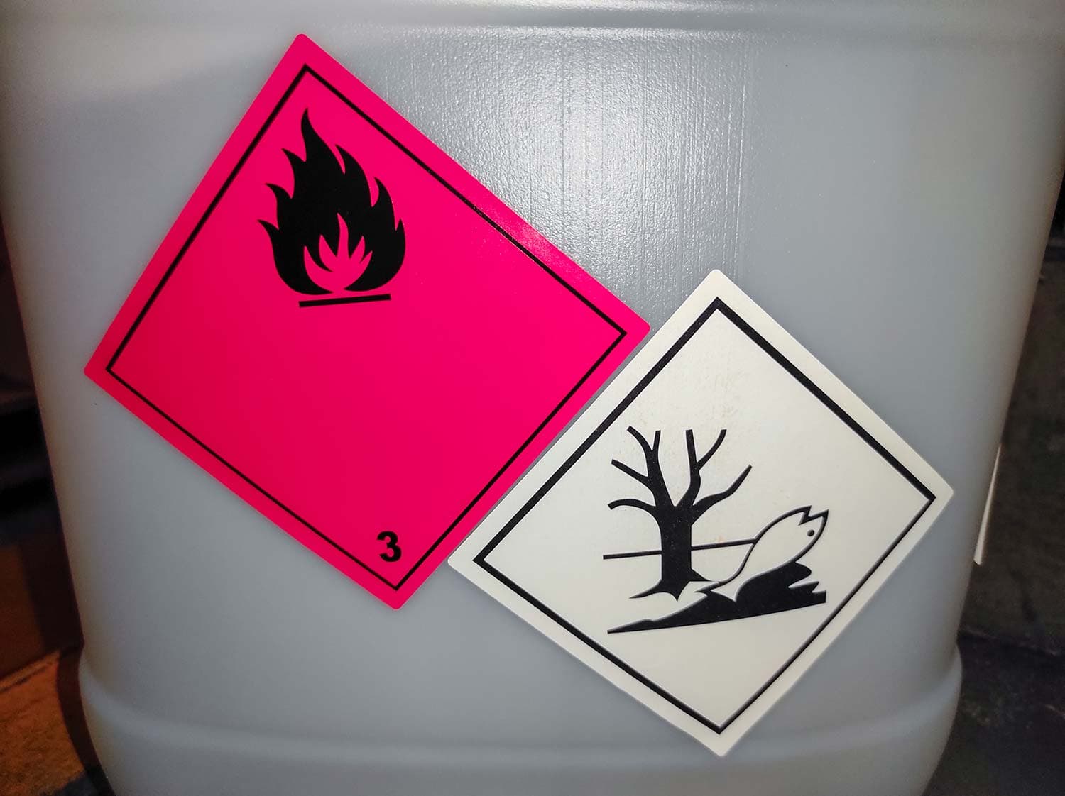 Gray plastic drum with a pink flammable class 3 placard and a white environmental hazard label showing a dead tree and fish