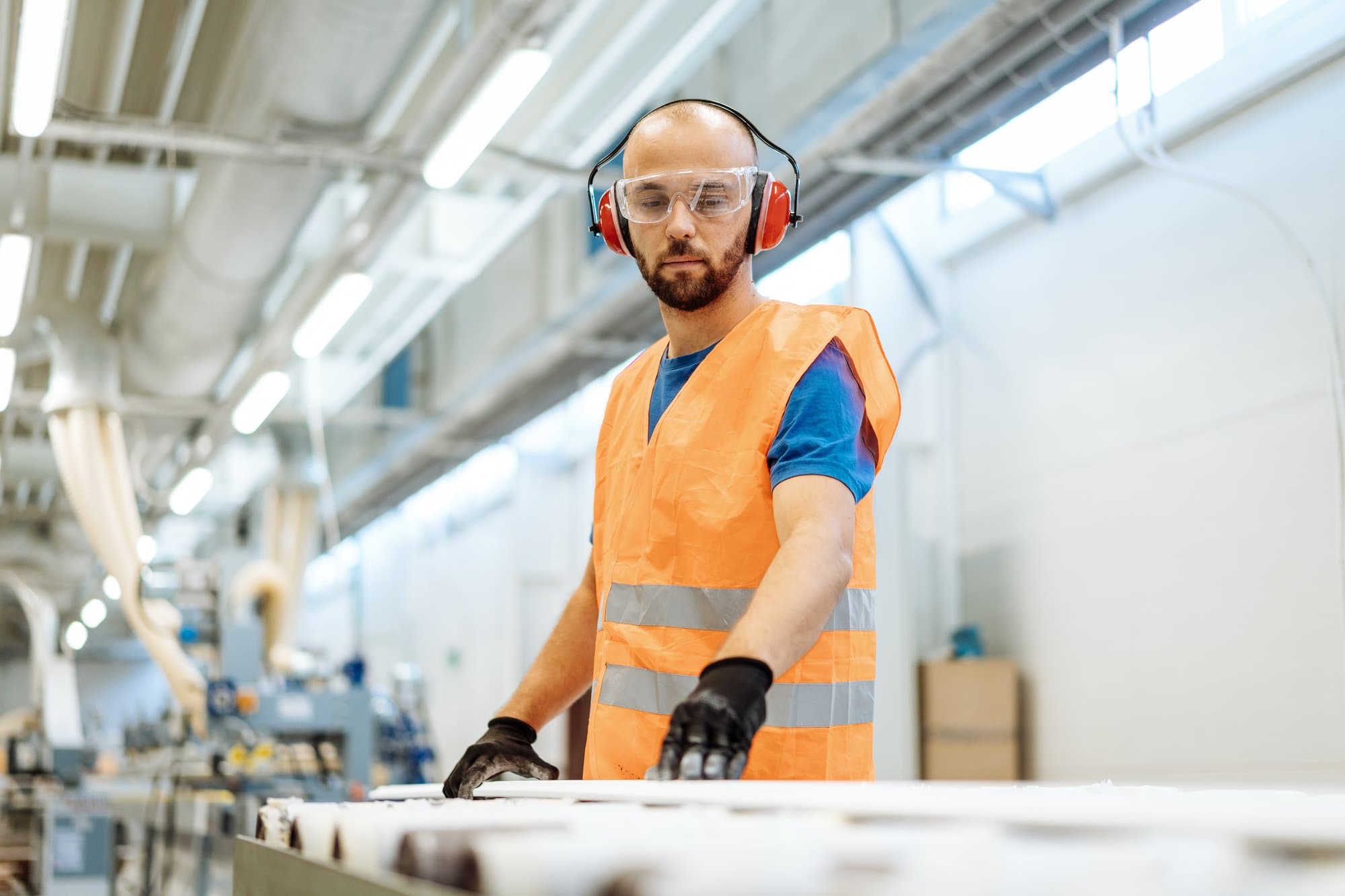 Worker wearing earmuffs, safety glasses, and a high-visibility vest in an industrial production facility.