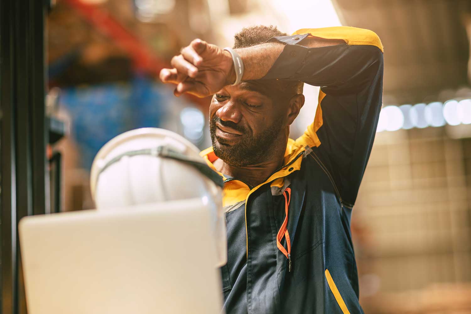 Warehouse worker in a navy and yellow jacket wiping sweat from his forehead with his forearm, white hard hat and laptop in front of him
