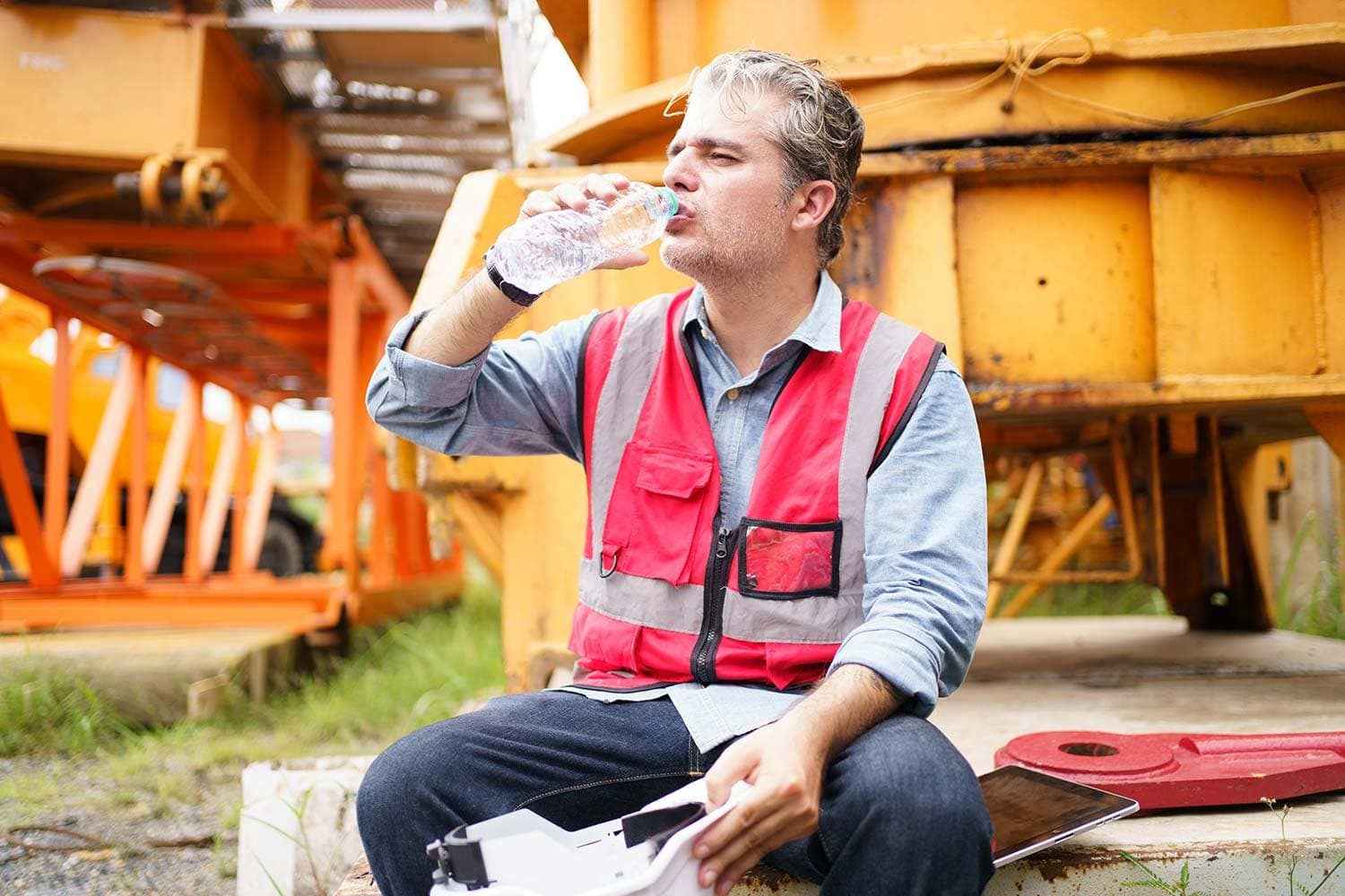 Worker in a red safety vest sitting beside yellow heavy equipment outdoors, drinking from a water bottle with a hard hat in his lap