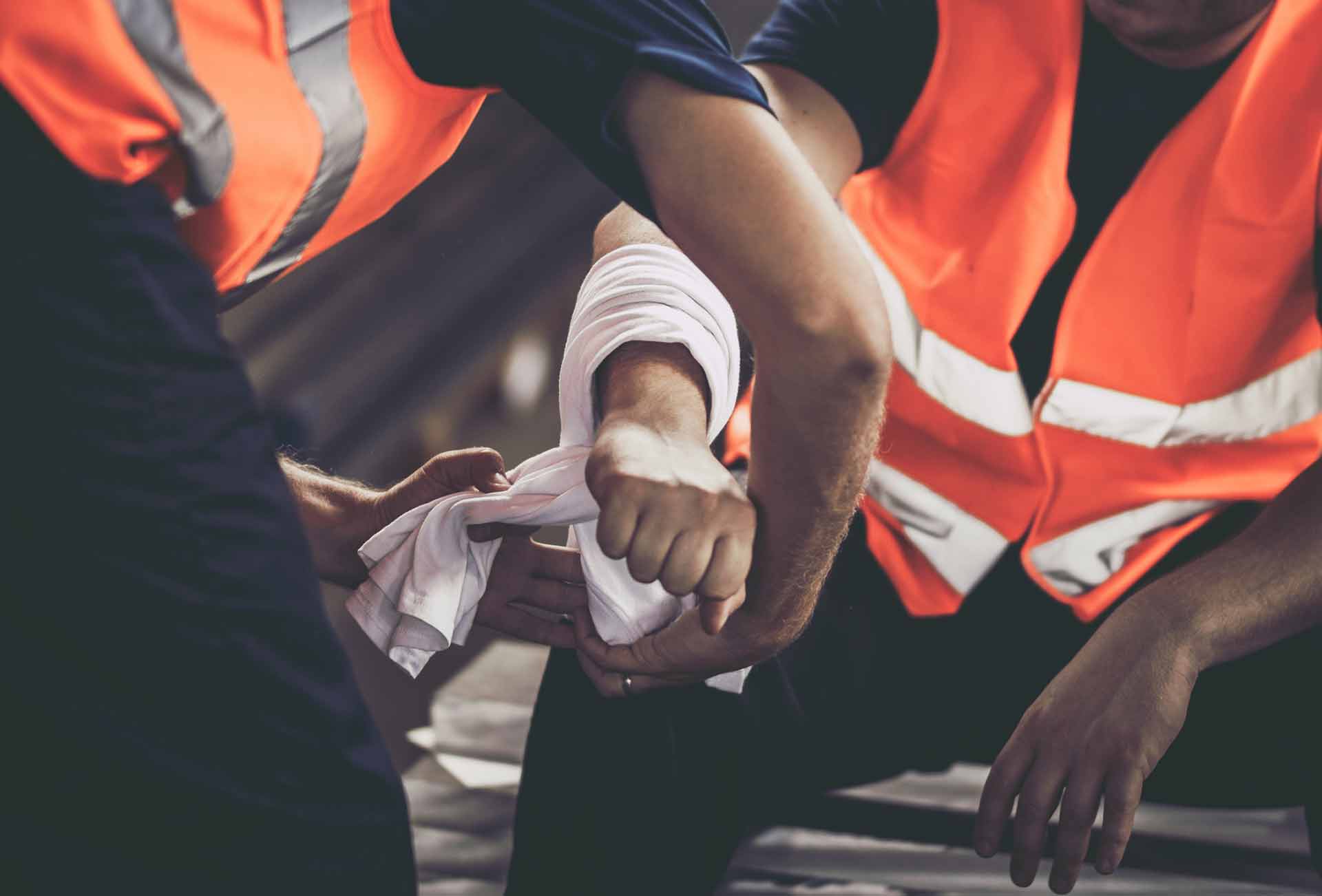 Workers in orange high-visibility vests wrapping a white cloth bandage around a coworker's injured forearm