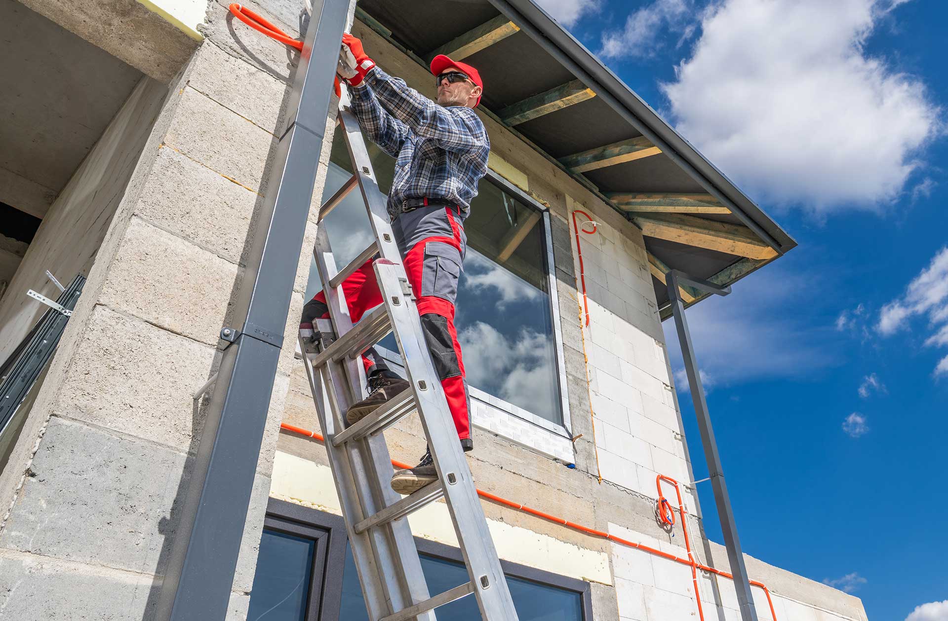 Worker in a red cap and plaid shirt standing on an aluminum extension ladder against a house under construction, blue sky behind