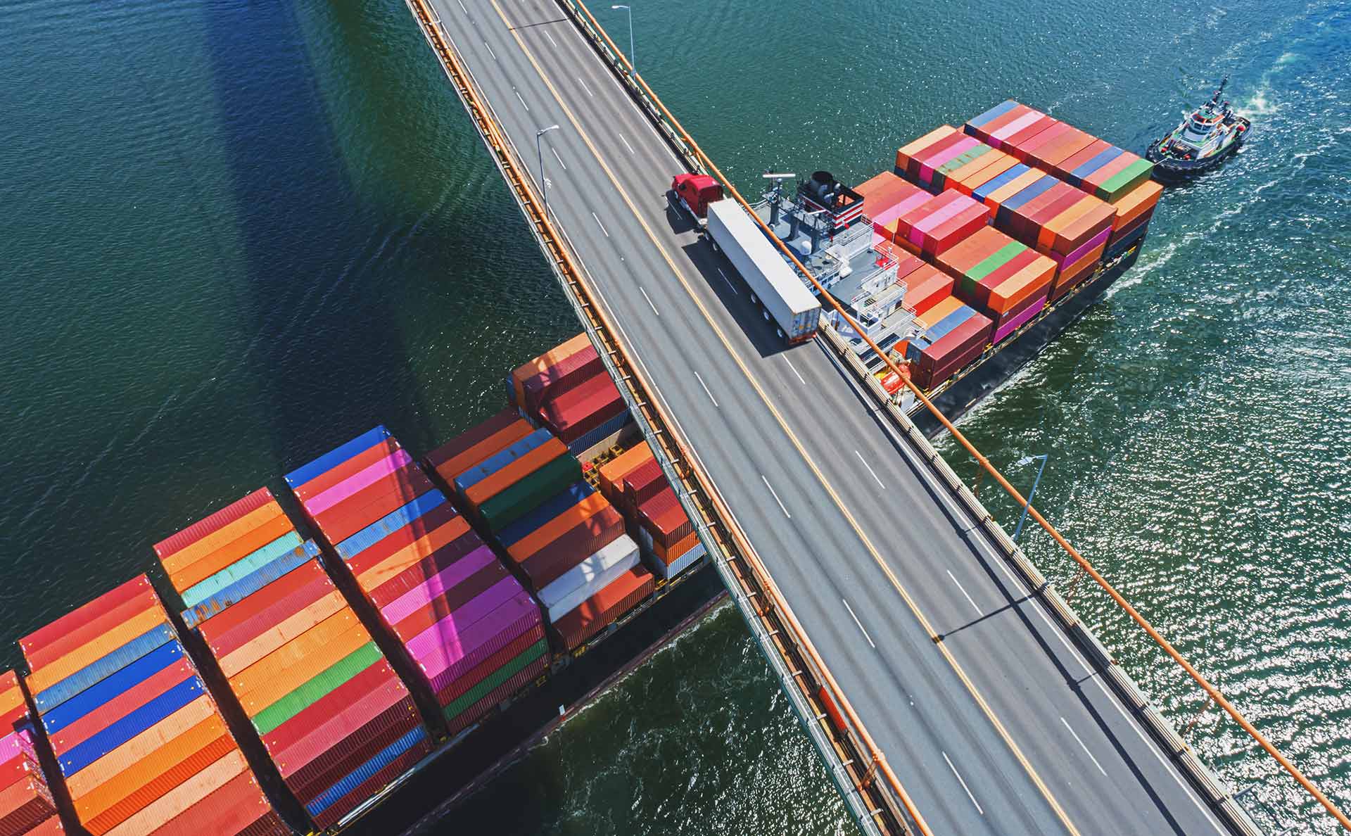 Aerial view of a semi truck crossing a bridge over water while barges stacked with colorful shipping containers pass below with a tugboat