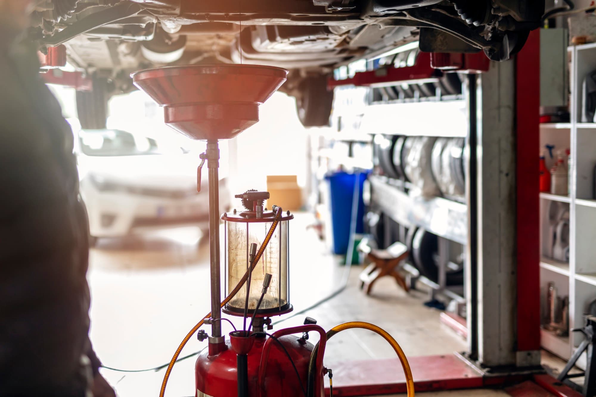 A red oil drain unit with a wide funnel and a fluid-filled sight chamber stands beneath a vehicle raised on a lift in an auto repair shop, with tires and parts shelving in the background.