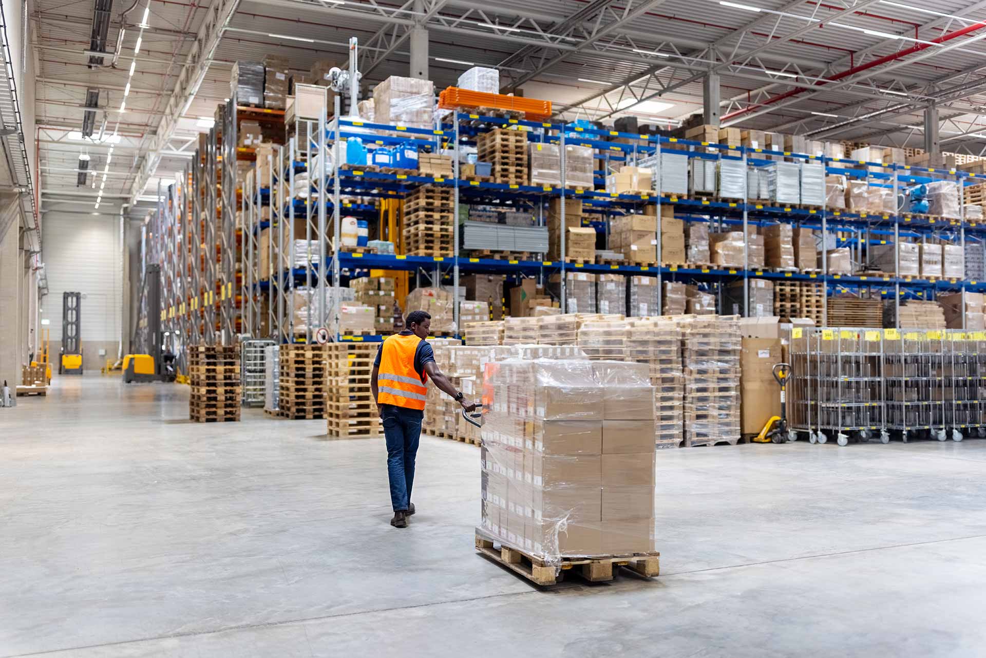 Worker in an orange safety vest pulling a pallet jack loaded with shrink-wrapped boxes through a large distribution warehouse