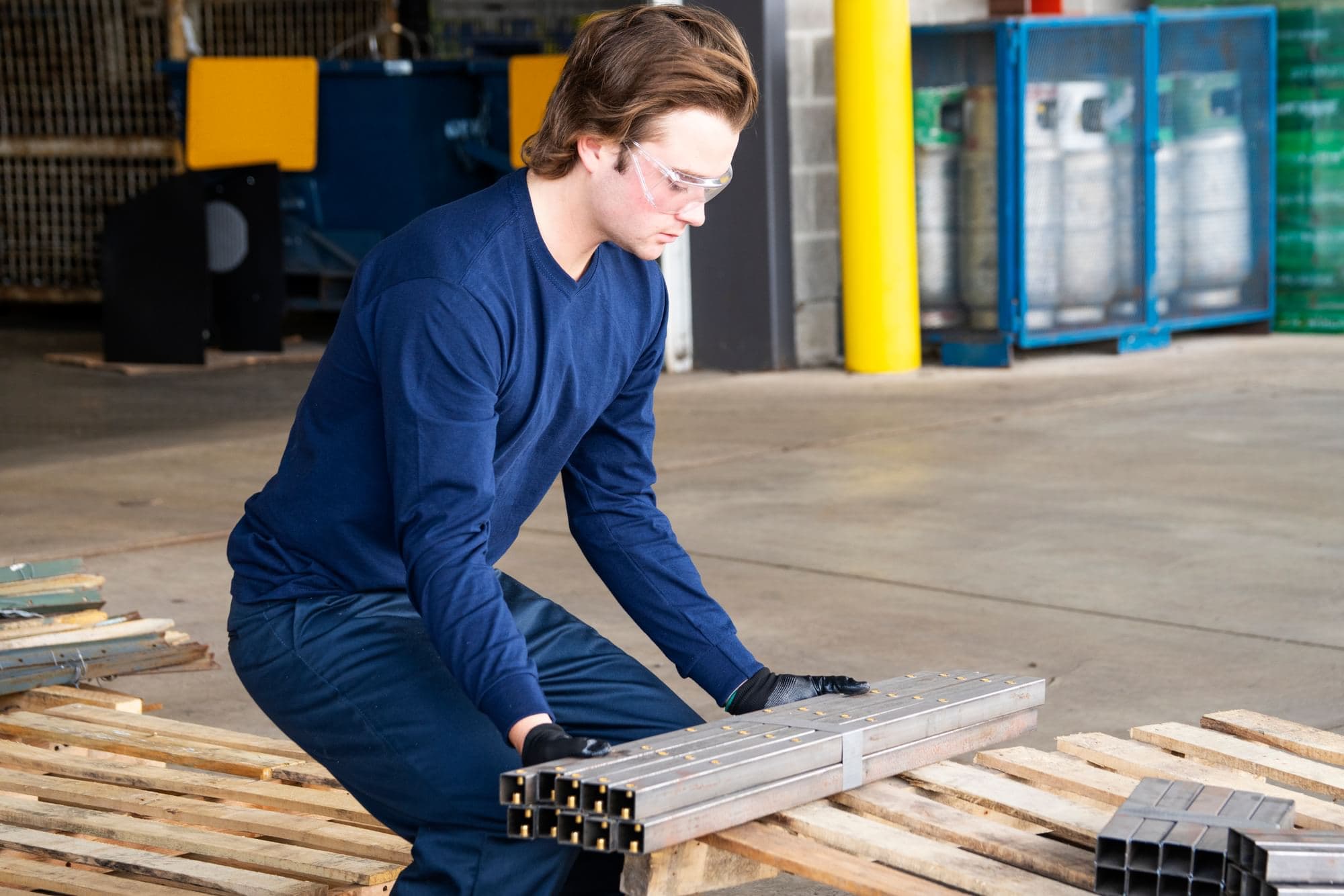 Worker wearing safety glasses and gloves handling a bundle of square metal tubing on a wooden pallet in an industrial work area.
