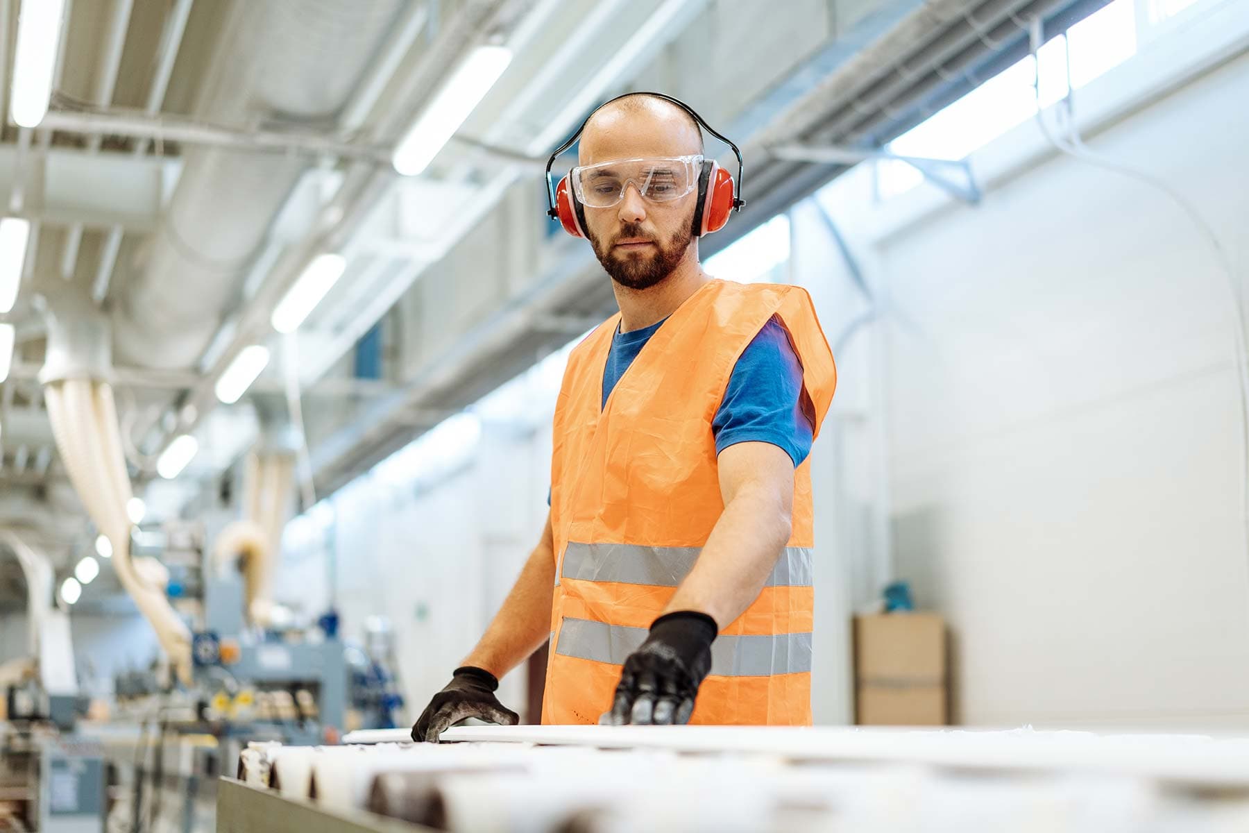 Worker wearing red earmuffs, safety glasses and an orange hi-vis vest handling material on a roller line in a bright production hall
