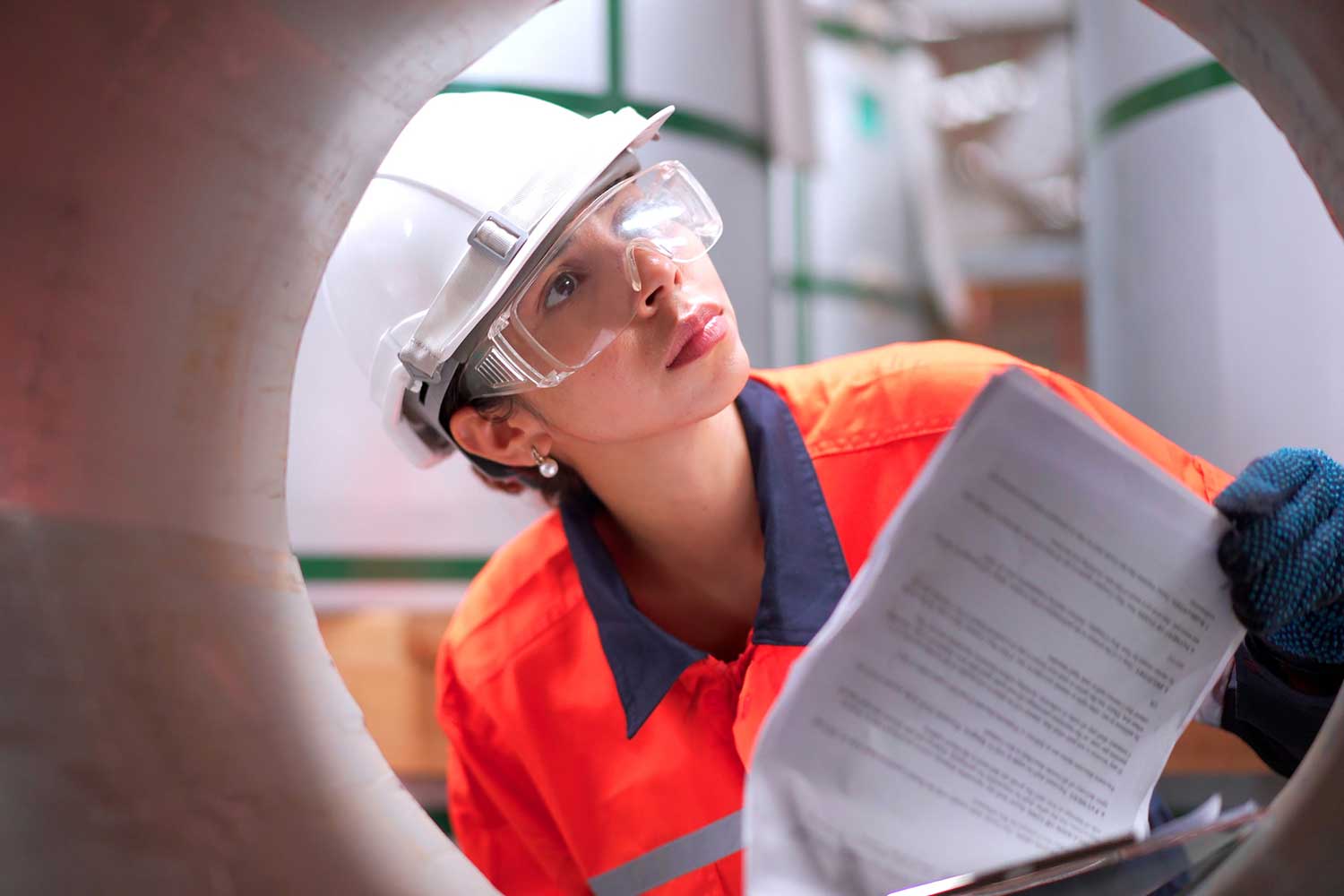 Woman in a white hard hat, safety glasses and orange workwear reviewing paperwork while inspecting the opening of a large steel pipe