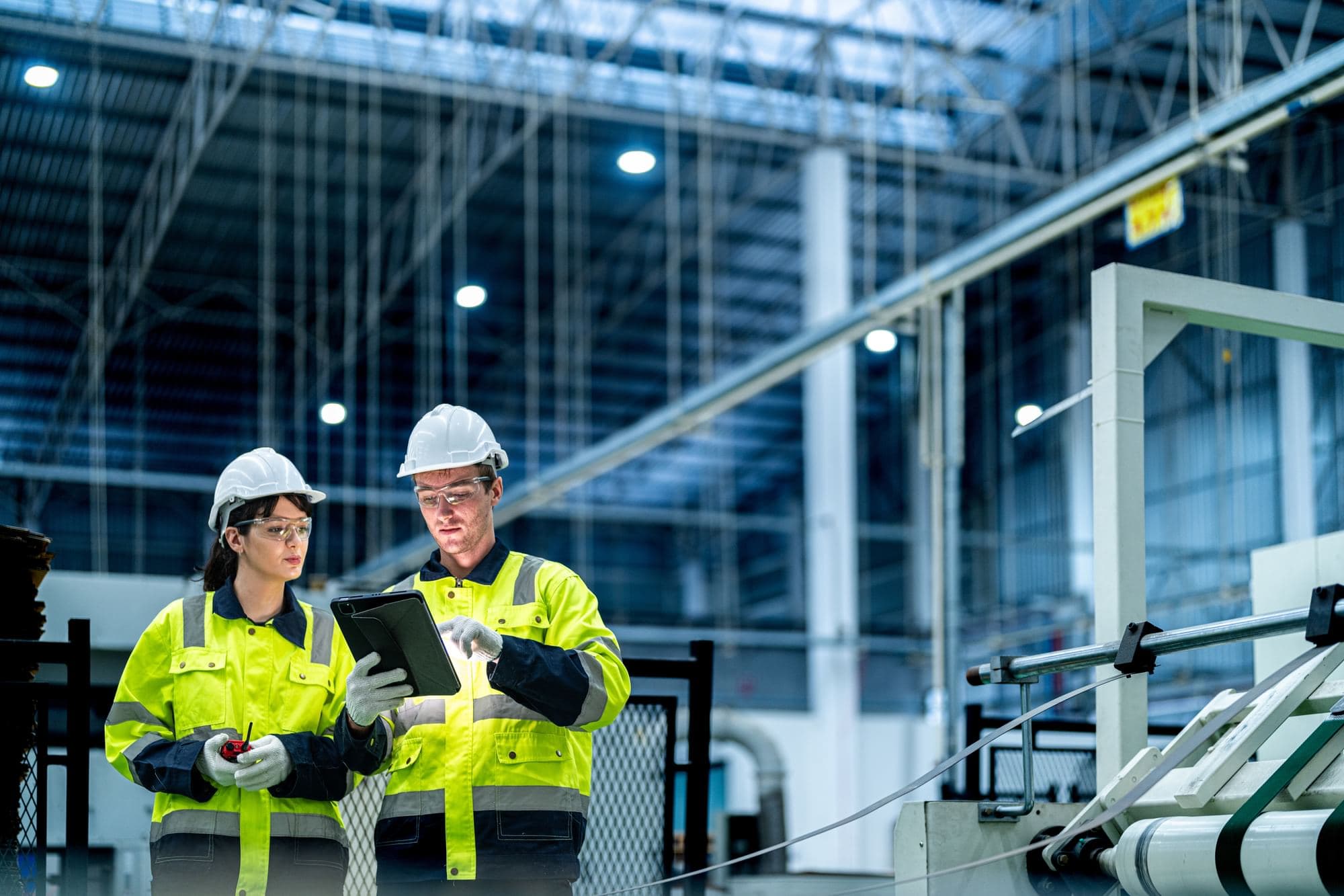 Two industrial workers wearing hard hats and high-visibility jackets reviewing information on a tablet.