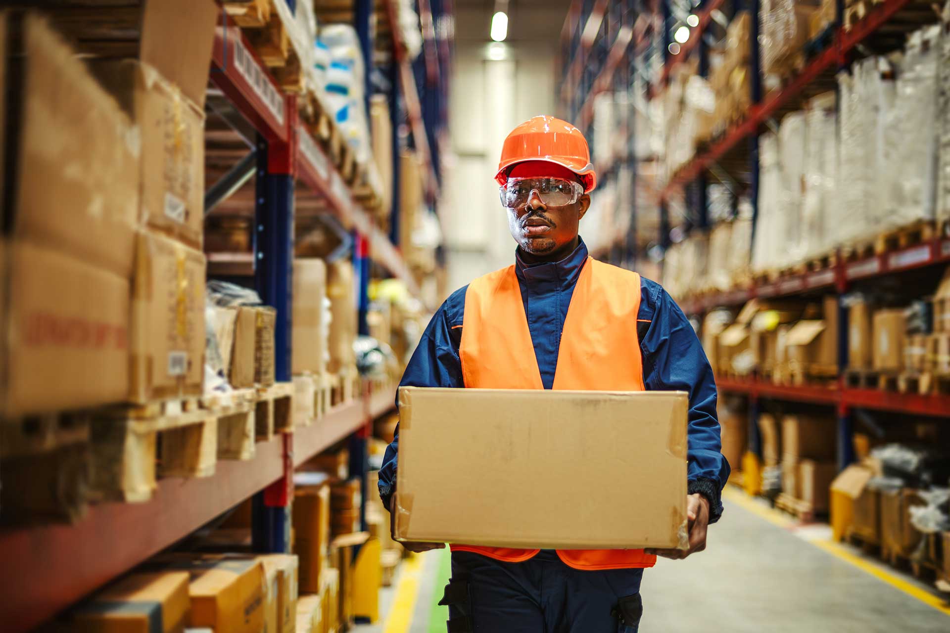 Warehouse worker in an orange hard hat, safety glasses and hi-vis vest carrying a cardboard box down a racking aisle