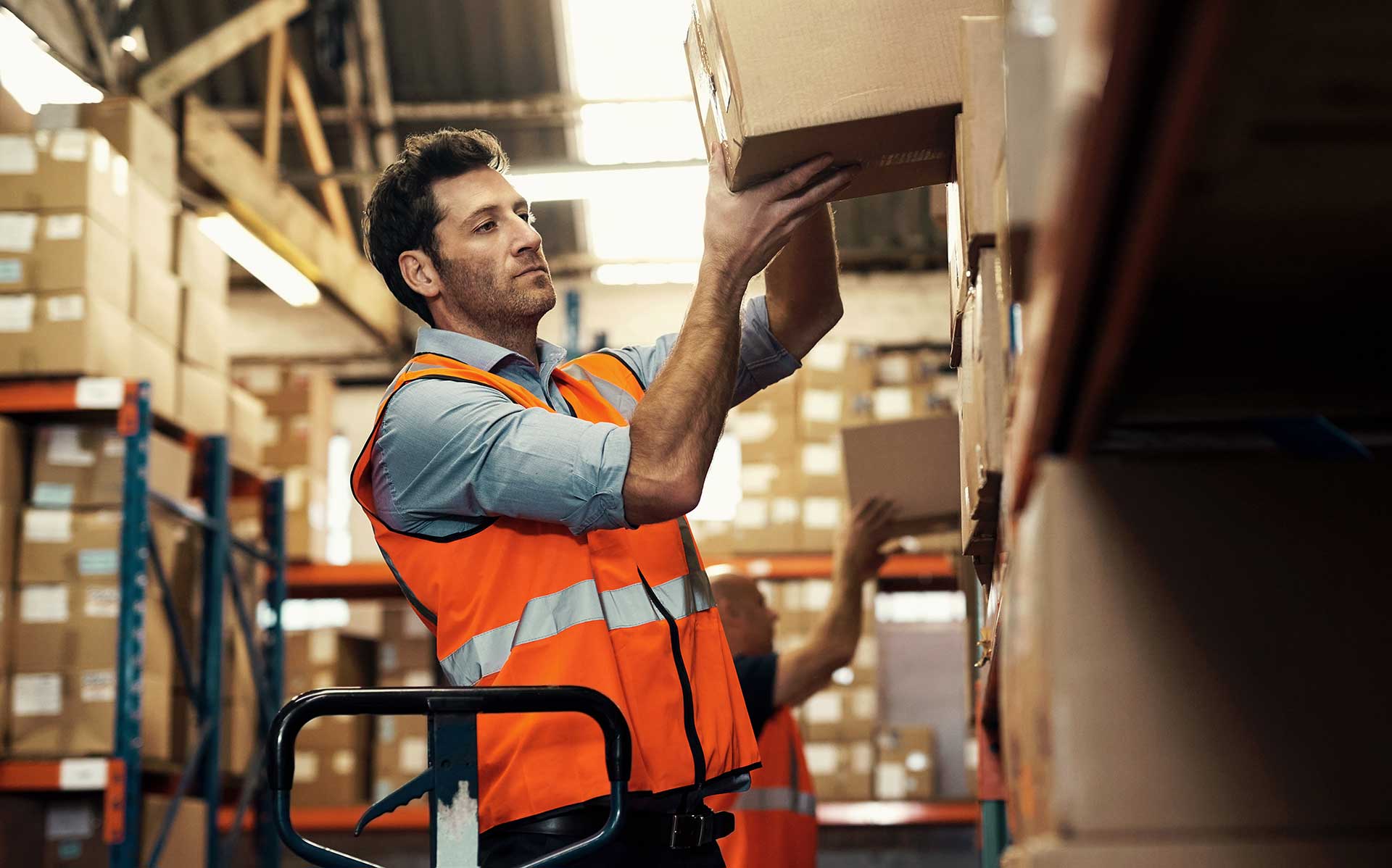 Warehouse worker in an orange hi-vis vest lifting a cardboard box onto a high shelf, with a pallet jack handle in the foreground