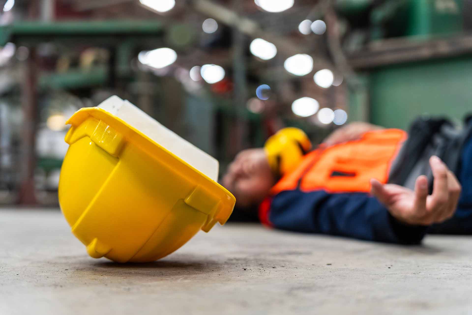 Yellow hard hat lying on a concrete factory floor with a fallen worker in an orange hi-vis vest blurred in the background