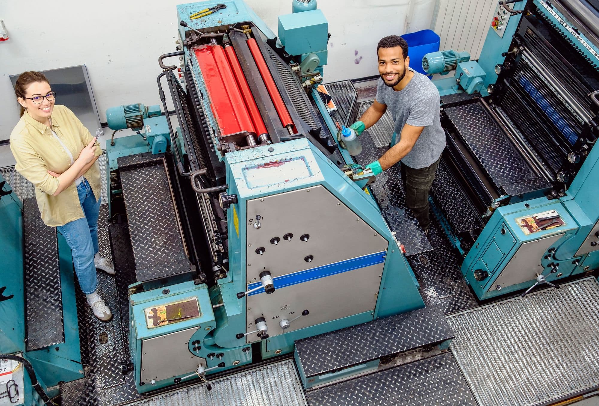 Two workers standing beside a large industrial printing press in a print shop.