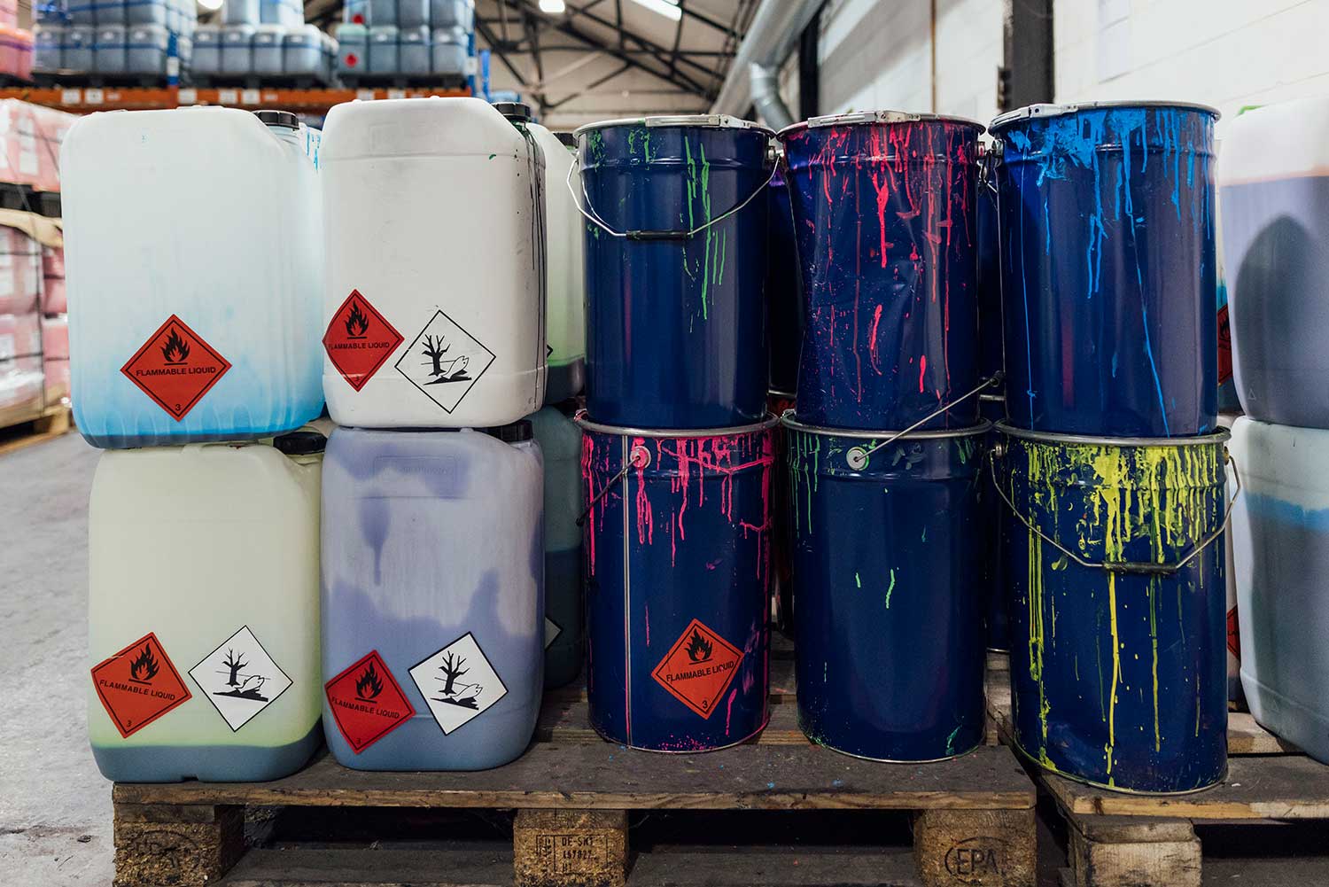 Plastic containers with flammable liquid warning labels stacked on pallets beside paint-splattered metal pails in a warehouse