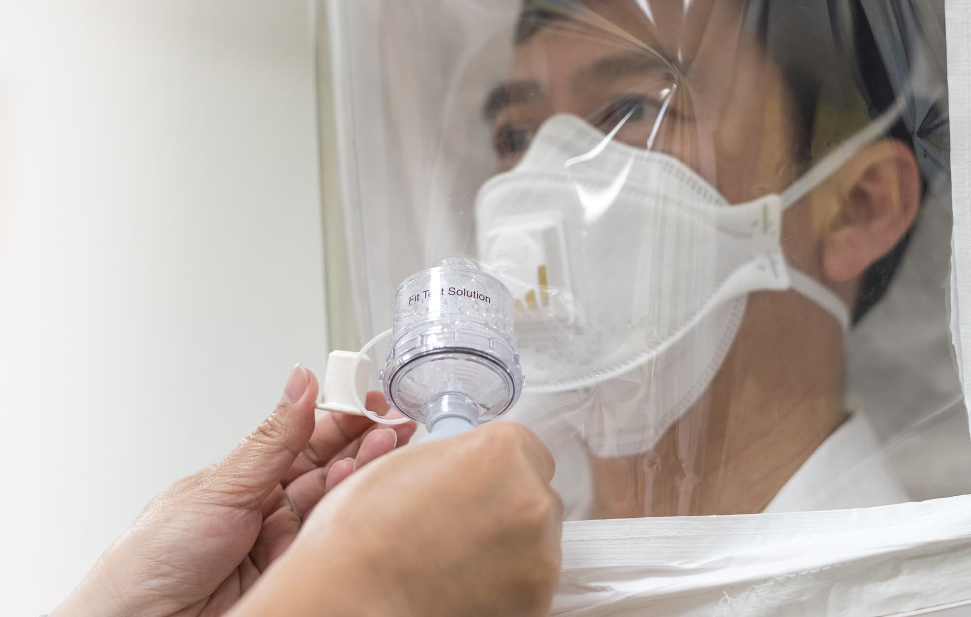 Man wearing a filtering facepiece respirator inside a clear fit-test hood as a tester holds a nebulizer labeled Fit Test Solution
