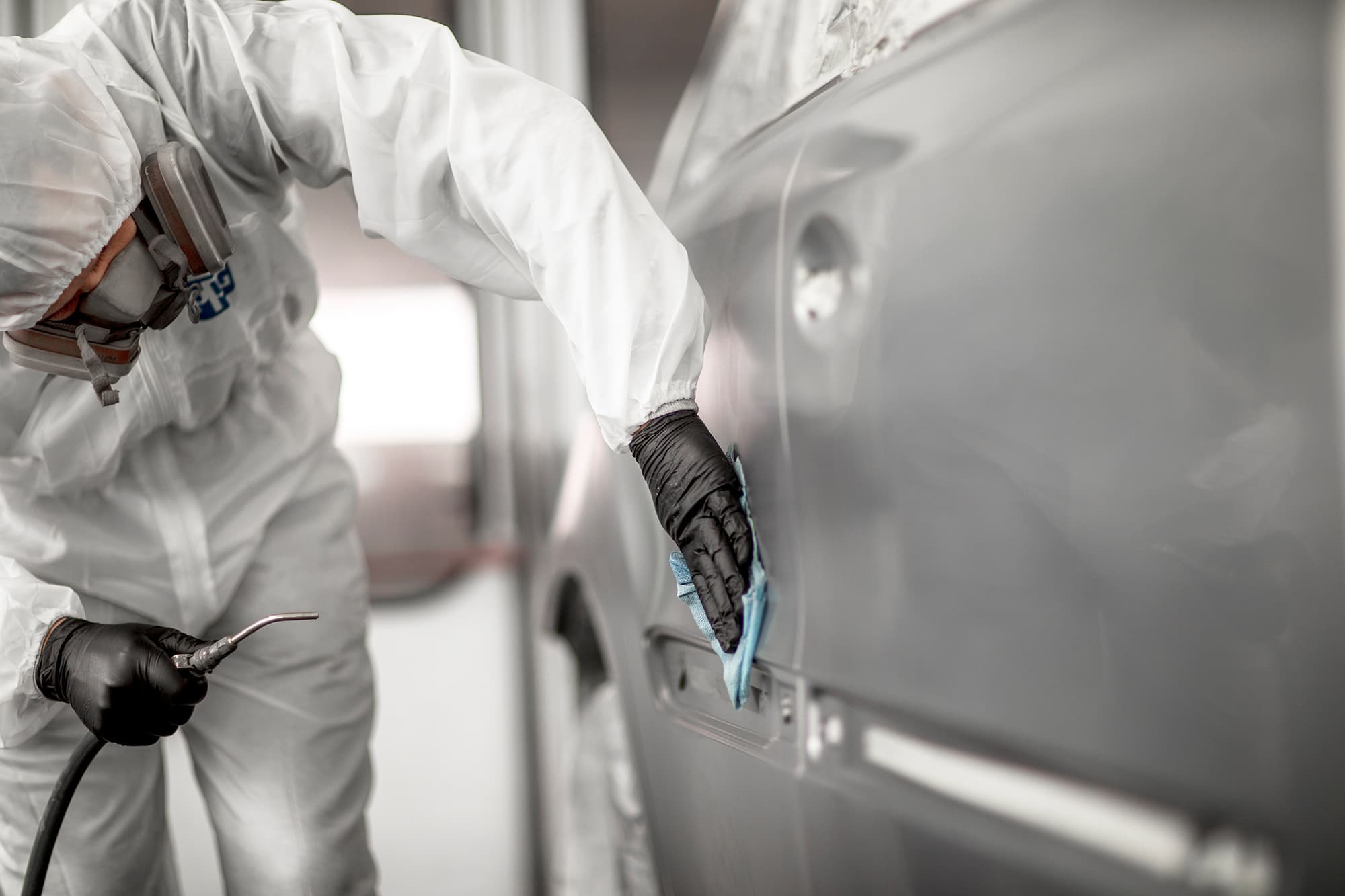 Painter in white coveralls, half-mask respirator and black gloves wiping a car door panel with a blue cloth in a paint booth