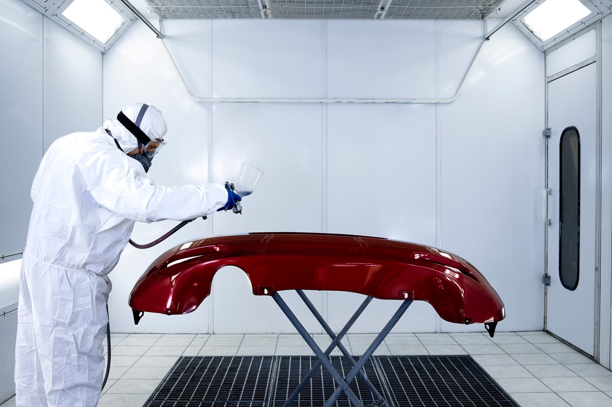 Painter in a hooded protective suit and respirator spray painting a dark red car bumper on a stand in a white spray booth