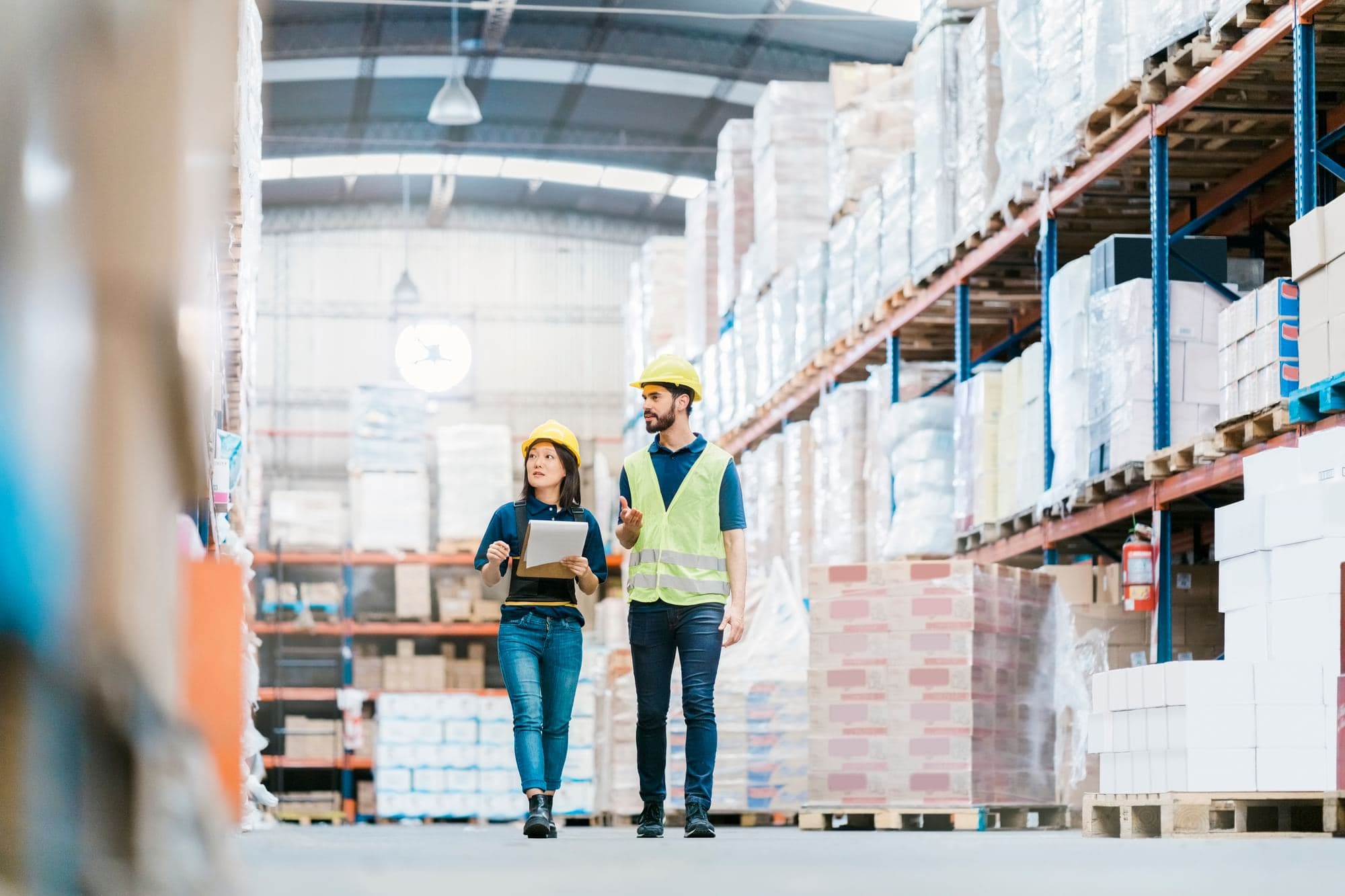 Two workers wearing hard hats walking through a warehouse aisle, with one holding paperwork.