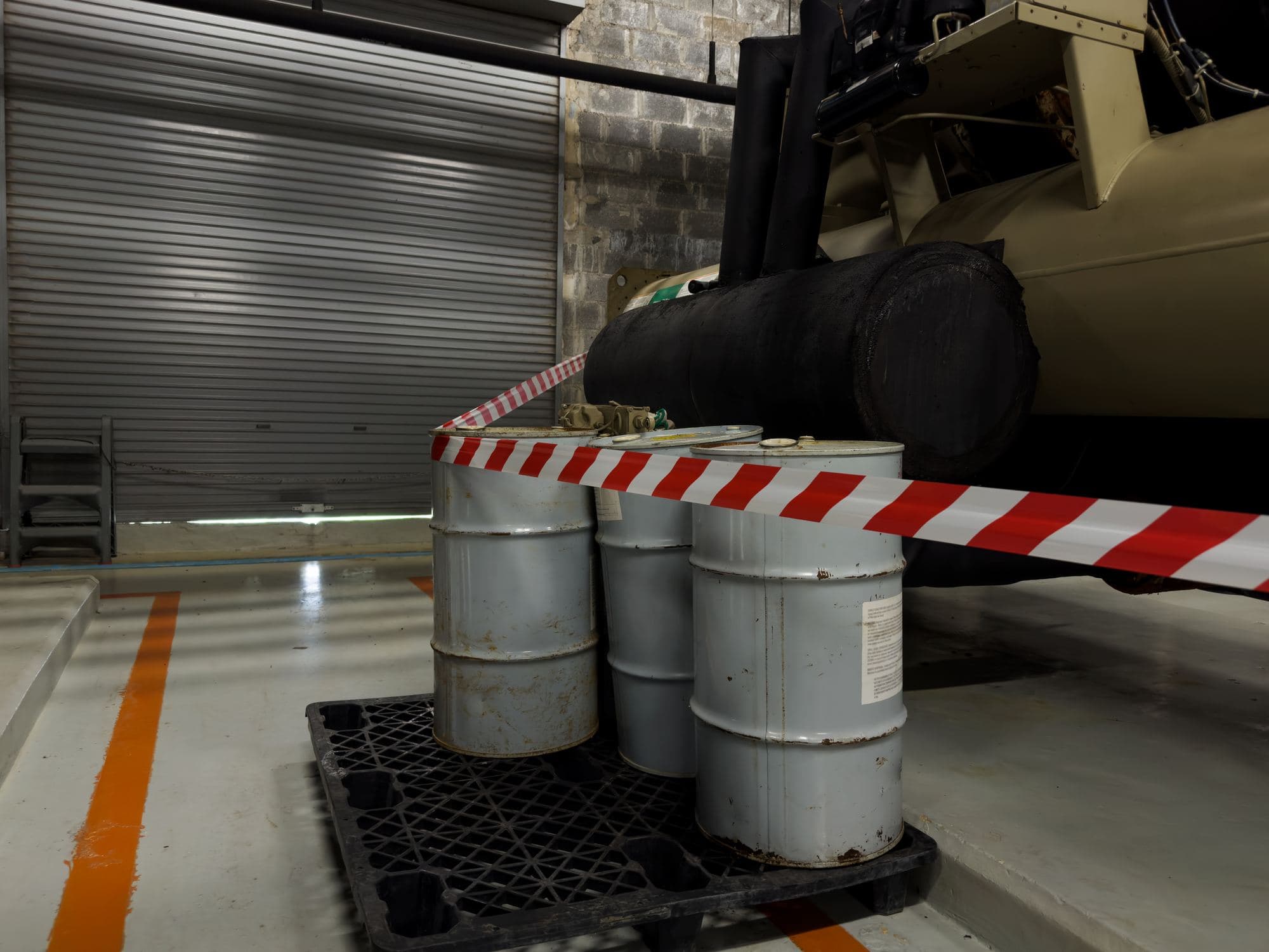 Metal drums stored on a spill-containment pallet beside industrial equipment, behind red-and-white barrier tape.