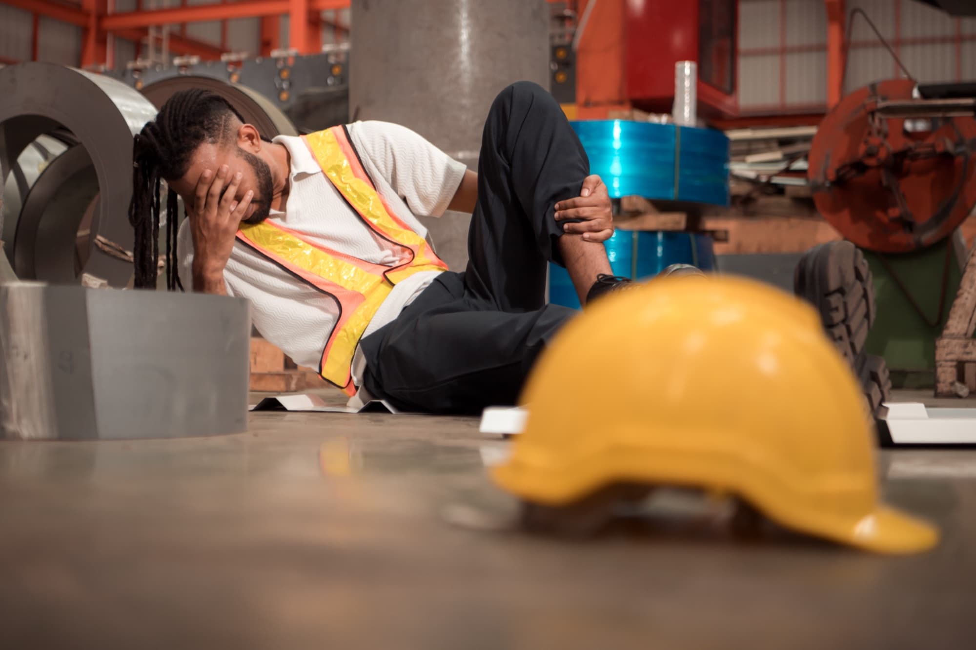Worker in a reflective safety vest sitting on the floor of an industrial facility, holding one hand to their face and the other to their lower leg, with a yellow hard hat on the floor in the foreground