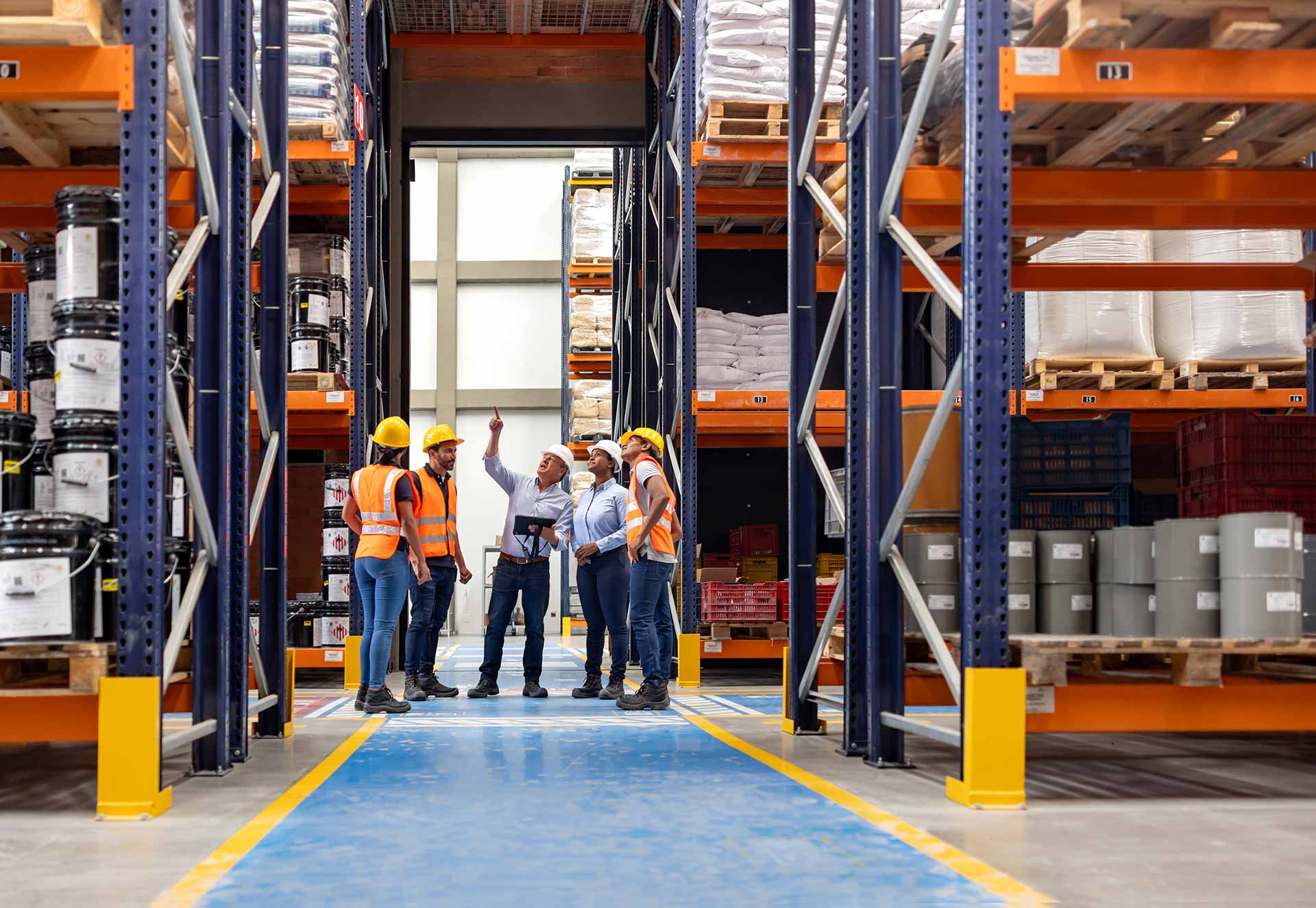 Five warehouse employees in hard hats and hi-vis vests gathered in a racking aisle while a supervisor points upward