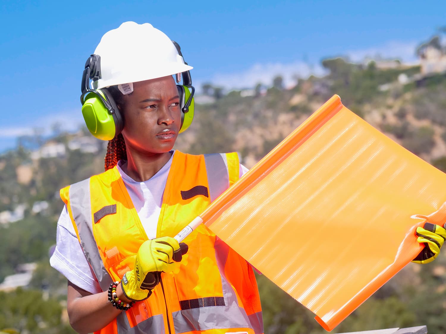 Flagger in a white hard hat with green earmuffs and an orange hi-vis vest holding a bright orange safety flag outdoors