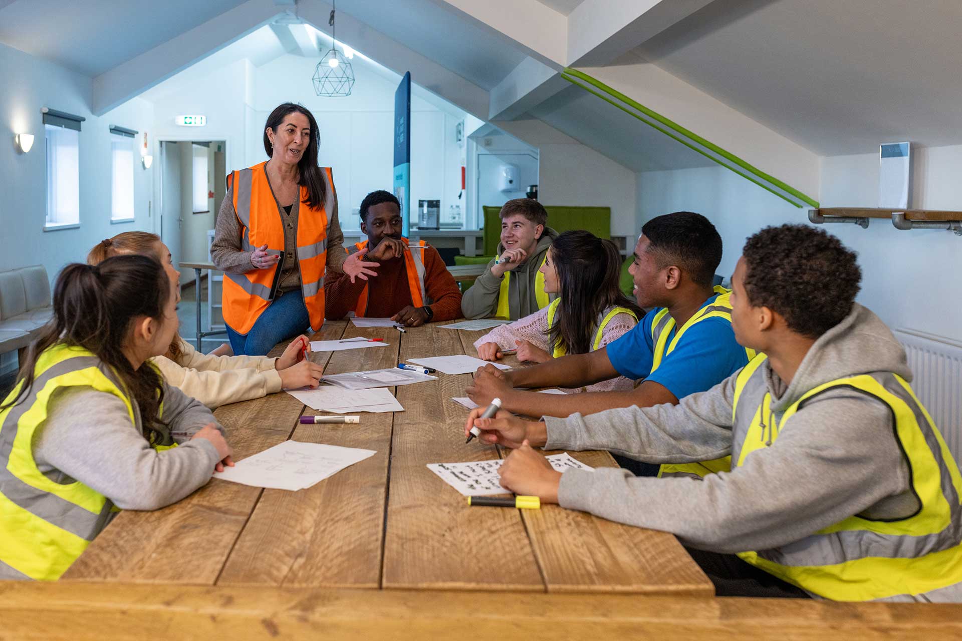 Instructor in an orange hi-vis vest leading a discussion with workers in yellow vests seated around a wooden table covered with papers
