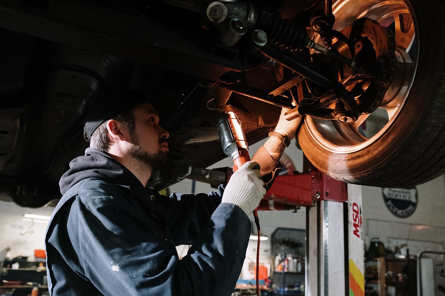 Mechanic in a black cap holding a work light to inspect the rear suspension and wheel of a car raised on a lift