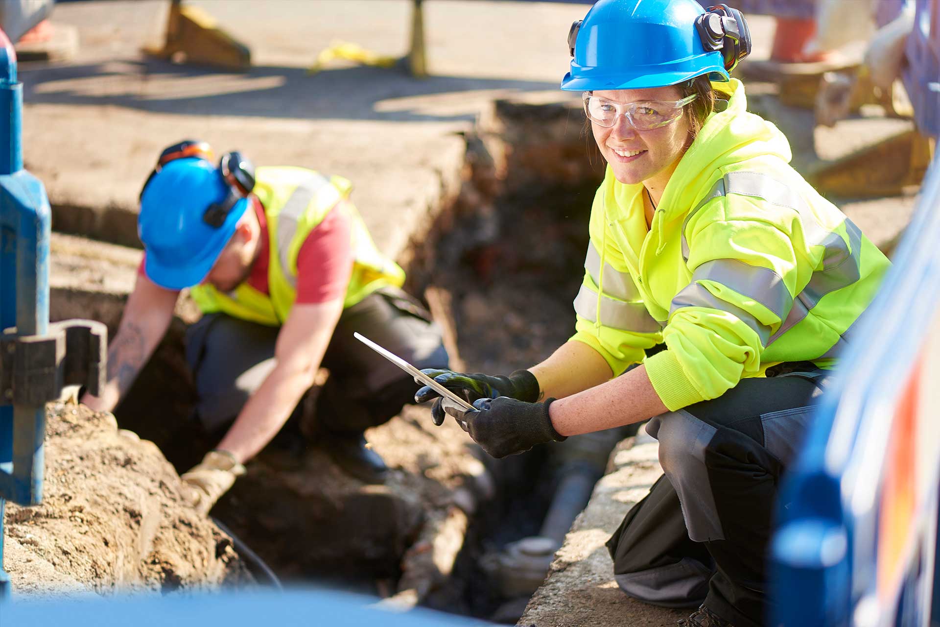 Worker in a blue hard hat and yellow hi-vis jacket holding a tablet at the edge of an open trench, with a coworker digging behind her