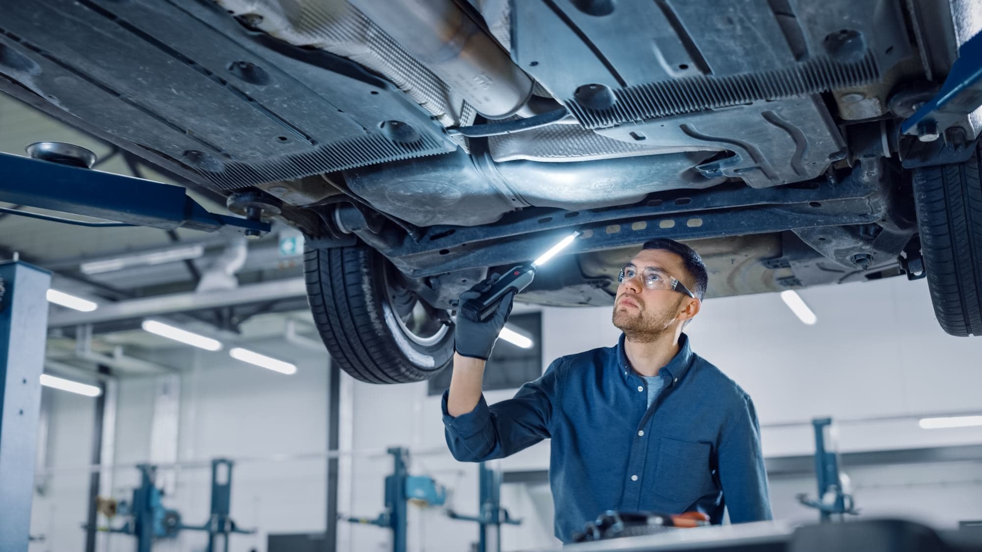 Automotive technician wearing safety glasses and gloves inspecting a vehicle underbody with a flashlight while the vehicle is raised on a two post lift
