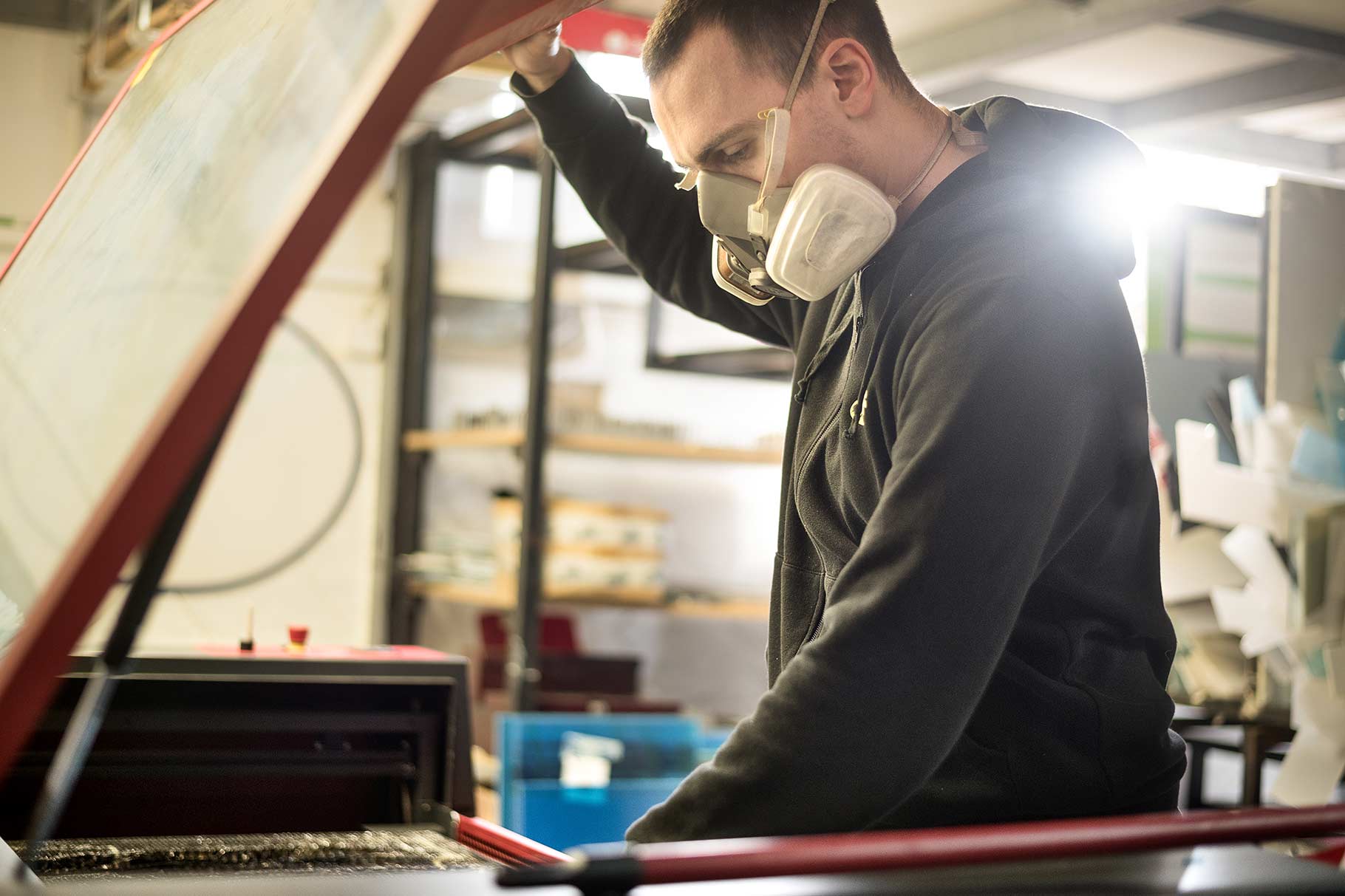 Print shop worker in a half-mask respirator lifting the hinged cover of a printing press to look at the bed below