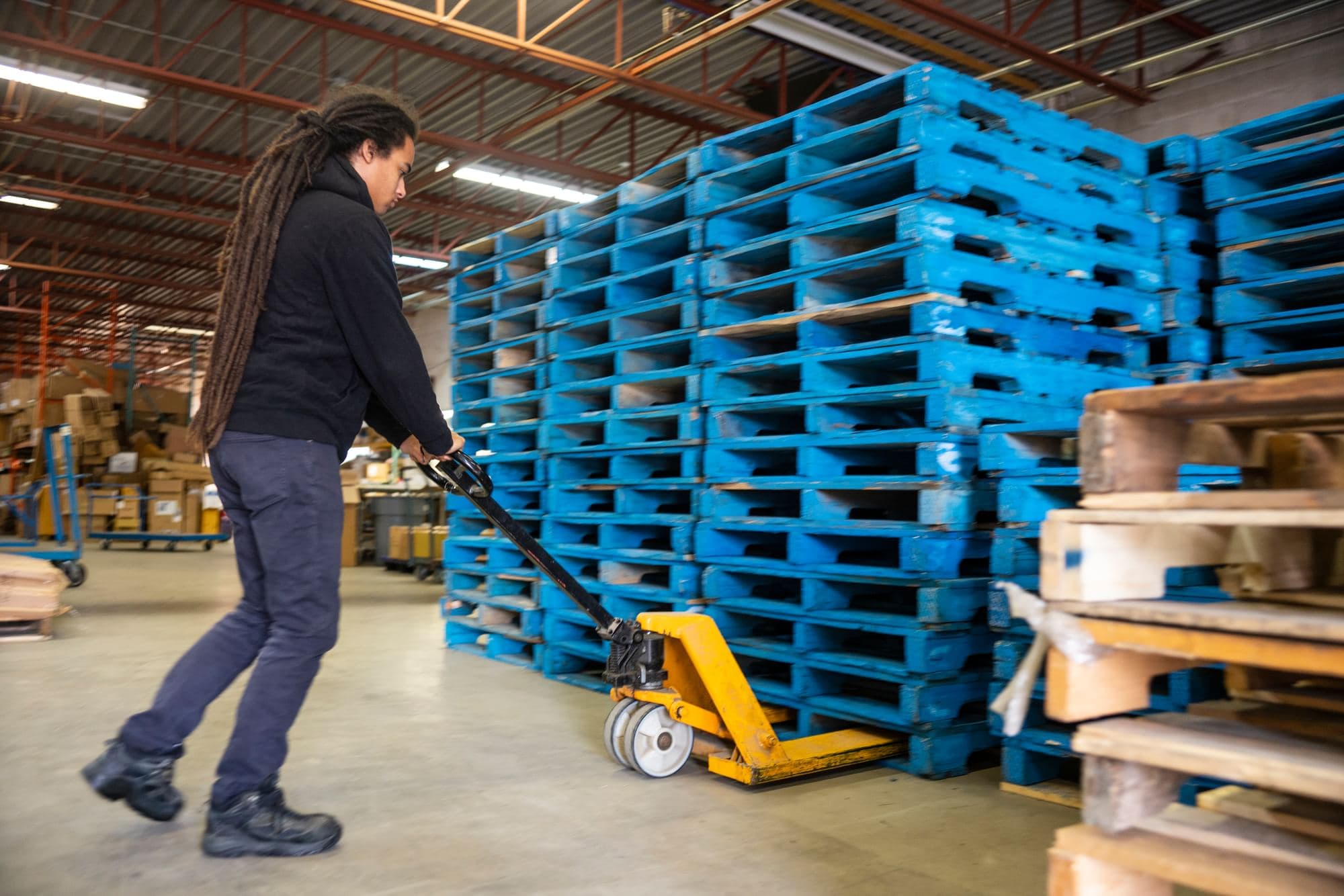 Worker using a manual pallet jack to move stacked blue pallets in a warehouse.